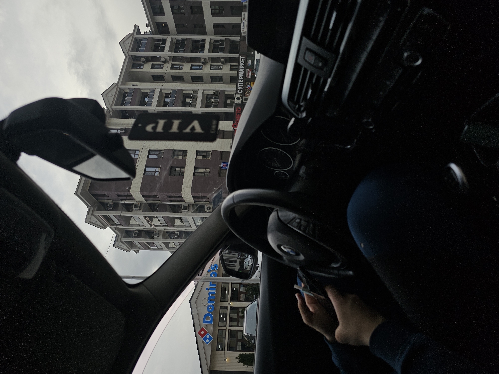 The image shows the view from inside a car, likely a BMW based on the steering wheel logo, in the city of Plovdiv, Bulgaria. The car is parked in front of a Domino's Pizza restaurant. Across the street, a tall, multi-story building with multiple windows and balconies is visible. A sign in Cyrillic indicates a supermarket, "СУПЕРМАРКЕТ", on the facade of the building. A "VIP" card or tag is hanging from the rearview mirror inside the car. A person's hand holding what appears to be a phone is visible to the right of the steering wheel. The sky is overcast.