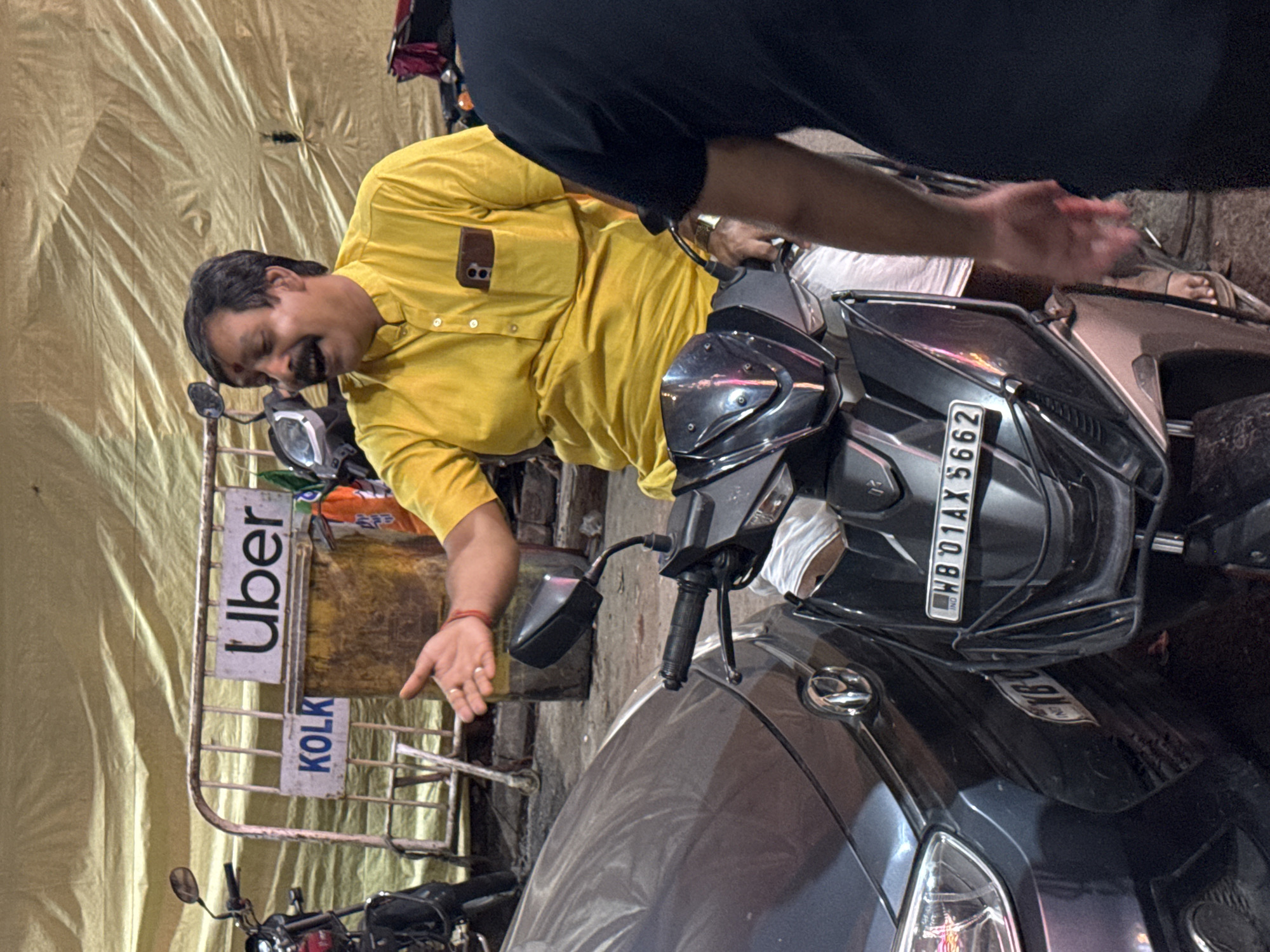 The image depicts a street scene in Kolkata, India, identifiable by the "Uber KOLK" sign, likely indicating an Uber pick-up/drop-off point. A man wearing a yellow shirt is standing on the side. A scooter is visible, its license plate reading "WB 01 AX 5662". A person wearing a black top with arms outstretched is also present. There is another person standing on the scooter. Other parked motorcycles are in the scene. The background shows a yellow sheet. The overall lighting suggests that this is an evening shot.