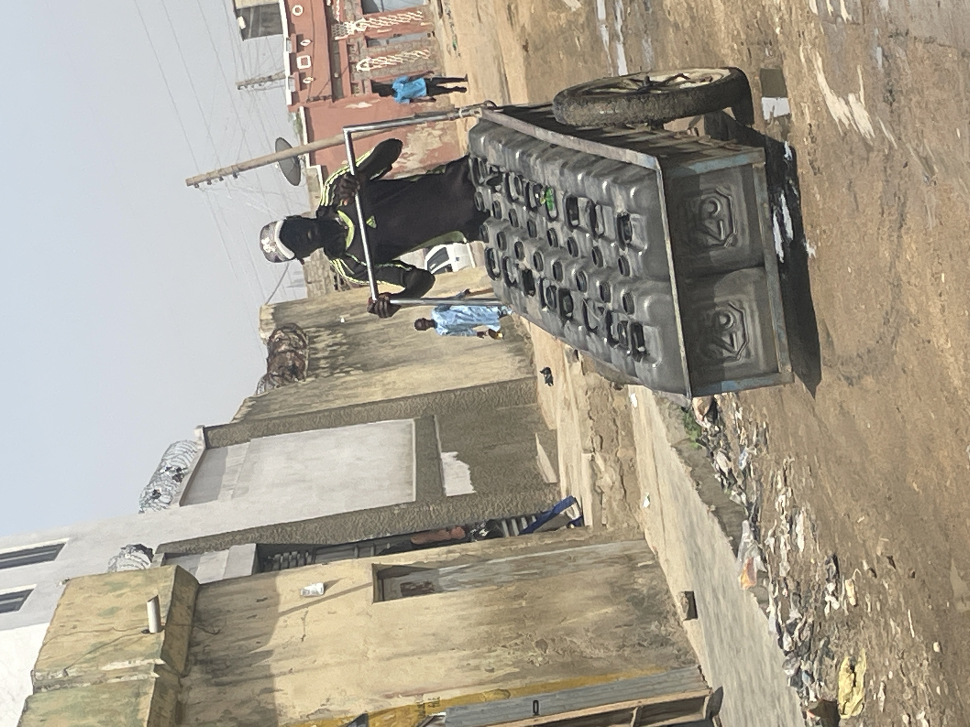 A man pulls a two-wheeled cart loaded with gray plastic containers along a dirt street in an urban area. He wears a black shirt with neon green stripes, dark shorts, a white helmet, and a face covering. Other people are seen in the background. The street has some trash along the side. The buildings in the background are primarily tan and gray, with some appearing to be in disrepair. Overhead electrical wires are visible. The scene appears to be in a sunny location.
