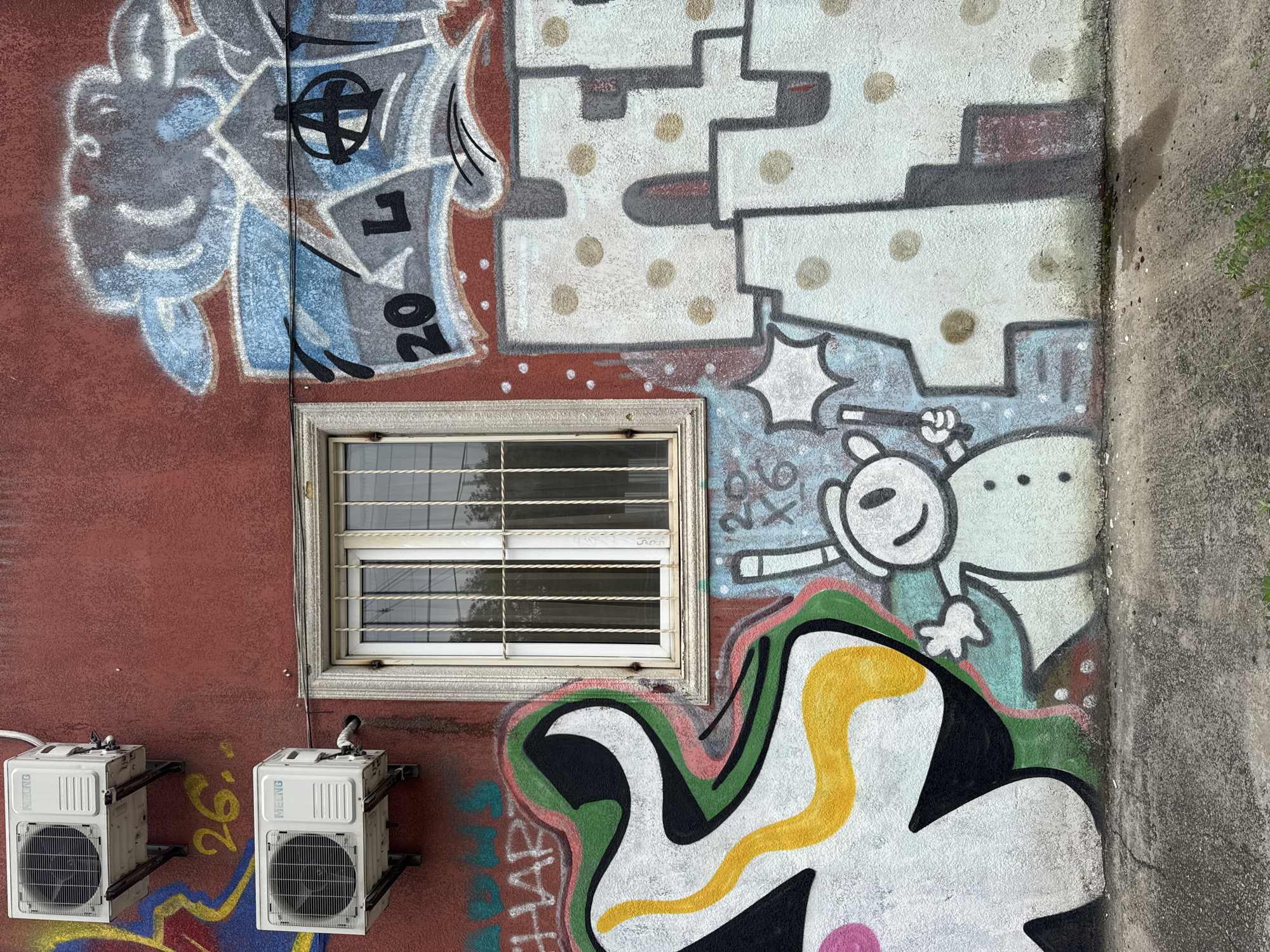 The image shows a section of an exterior wall covered in graffiti. Two white air conditioning units are mounted on the upper left of the wall. Below them, a window with metal bars is visible, set within a decorative stone frame. The wall itself is painted a dark red and is extensively decorated with various graffiti tags and drawings. On the left, a large, abstract shape in black, white, green, and yellow is depicted. To the right of the window, there is graffiti with blue, gray, and white tones, including the numbers "20" and the letter "L", along with a symbol resembling an anarchy sign. Below this, a cartoonish white figure with a black face, holding a stick, is drawn. Further to the right, large, blocky white letters with polka-dot-like patterns are visible, partially obscured by other graffiti. The ground in front of the wall is a cracked concrete surface with some small green plants growing. The scene is likely located in an urban environment, possibly a residential or commercial building, and the graffiti suggests a street art or urban culture context.