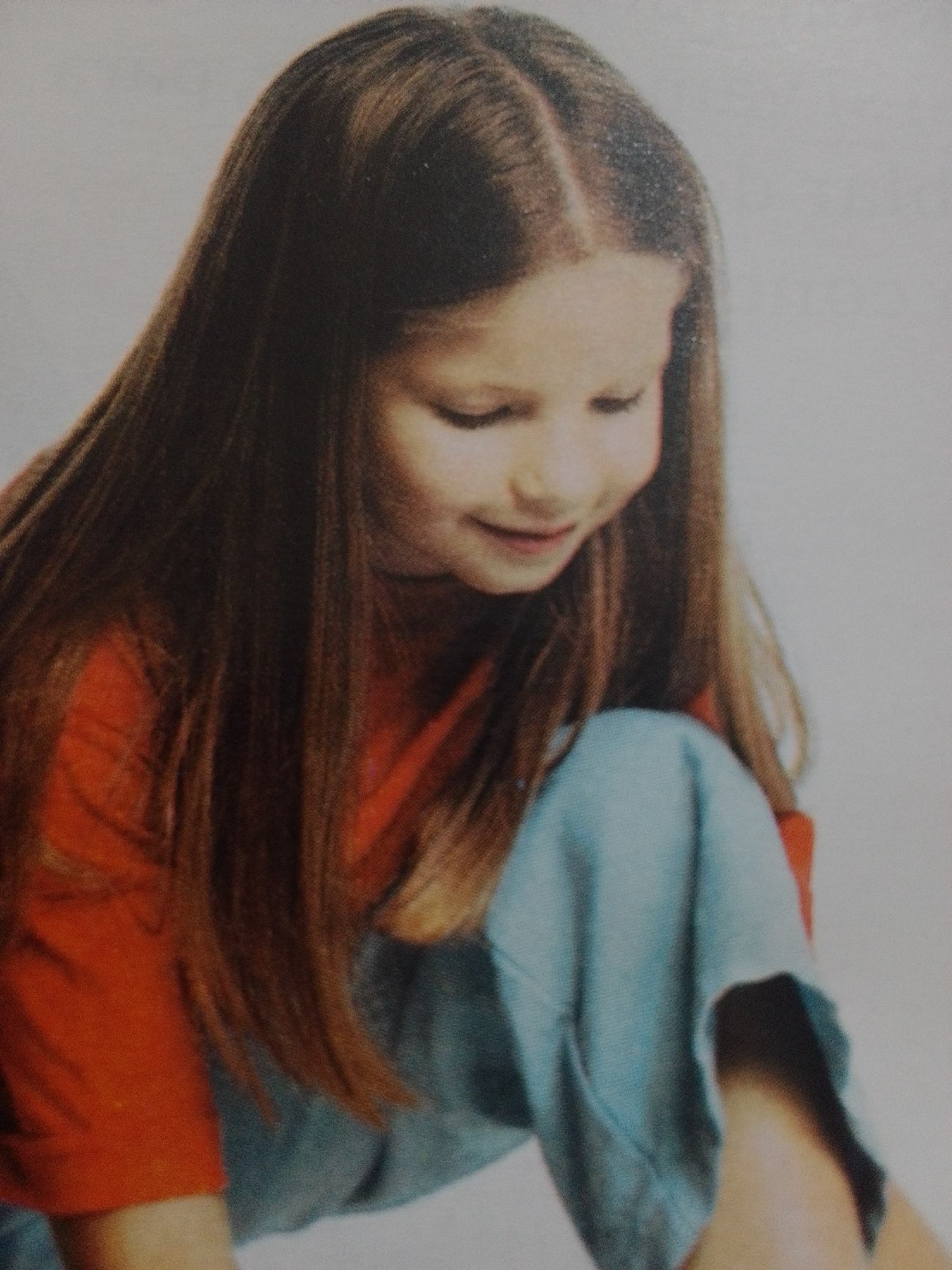 The media file is a medium shot, portrait-style photograph of a young girl. She has long, dark brown hair parted in the middle, framing her face. Her head is tilted downwards, and she is looking to her right with a gentle, closed-mouth smile. Her eyes are closed, and her eyebrows are slightly furrowed, suggesting a moment of quiet contemplation or perhaps a happy memory.
She is wearing a bright orange long-sleeved shirt and what appears to be light blue pants or a skirt. The background is a plain, light gray, providing no specific environmental context. The lighting is soft and diffused, illuminating her face from the front and slightly from the left. There are no discernible objects or other people in the frame.
The image has a vintage or slightly aged quality, possibly due to the printing process or the age of the photograph itself. The focus is on the girl's serene expression and her youthful appearance. There is no visible text within the image. The overall mood is peaceful and introspective. The photograph appears to be a personal portrait, capturing a candid moment.