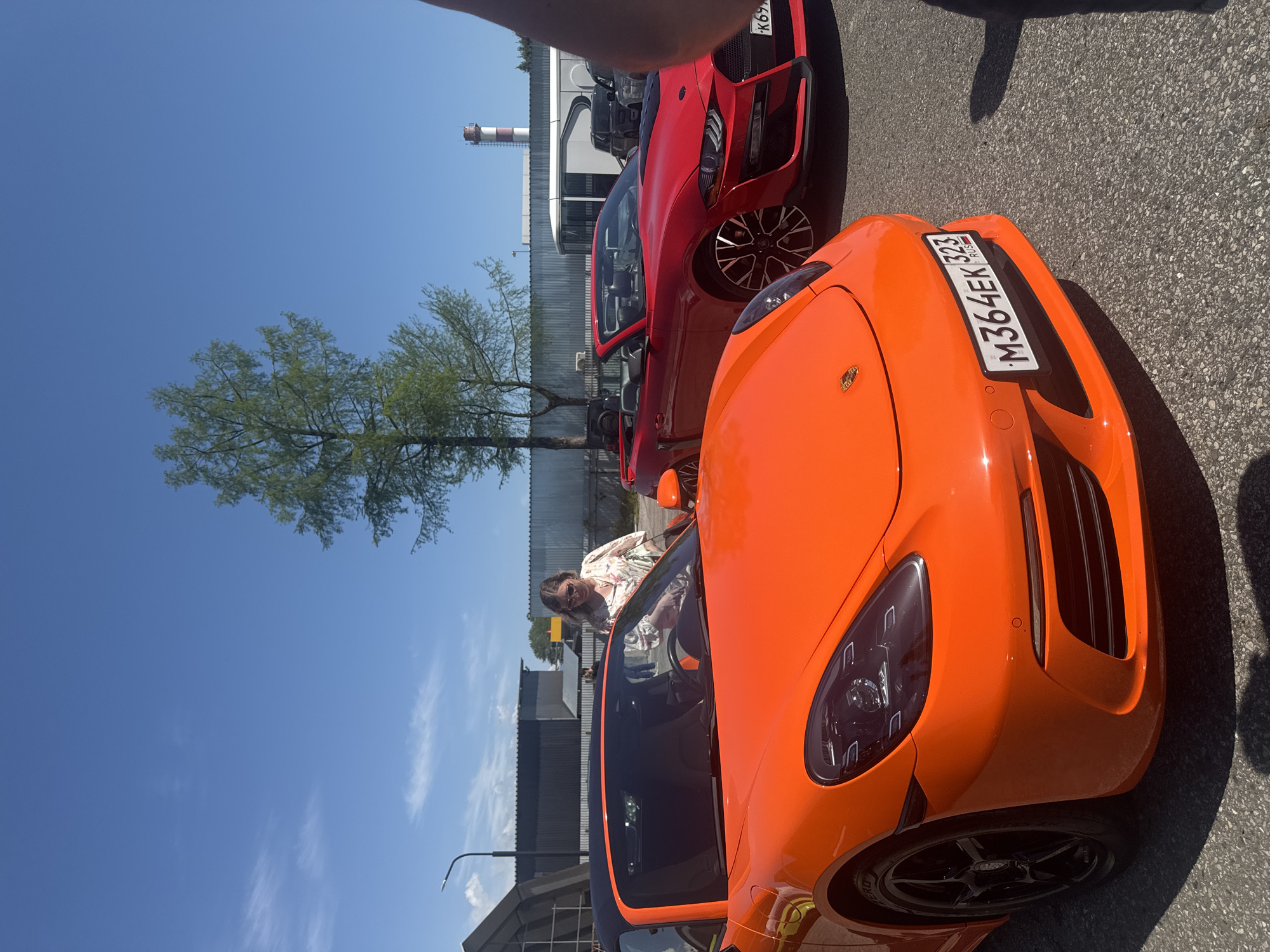 The image shows two cars, one orange and one red, parked on an asphalt surface. The orange car, a Porsche with a Russian license plate "M364EK 323", is in the foreground. A woman is seen inside the orange car. Behind it, a red car is partially visible. A tree and a smokestack are visible in the background. The setting appears to be a parking lot, possibly located in Sochi, Russia, based on the license plate.