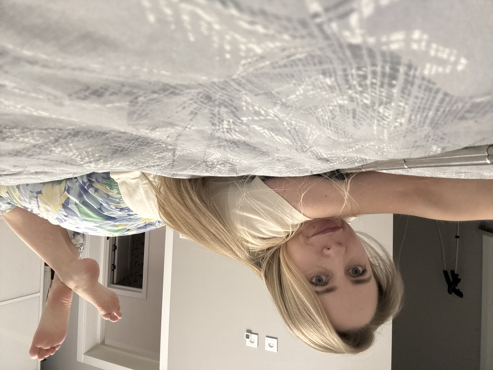 A blonde-haired woman with light skin lies in bed, looking at the camera. She wears a white t-shirt and floral-patterned pajama pants. Her bare feet are visible. A patterned bedsheet covers the majority of the top of the frame. In the background, a window and electrical outlets are visible. There is no explicit evidence to pinpoint the exact location as Chaikovsky, Russia.