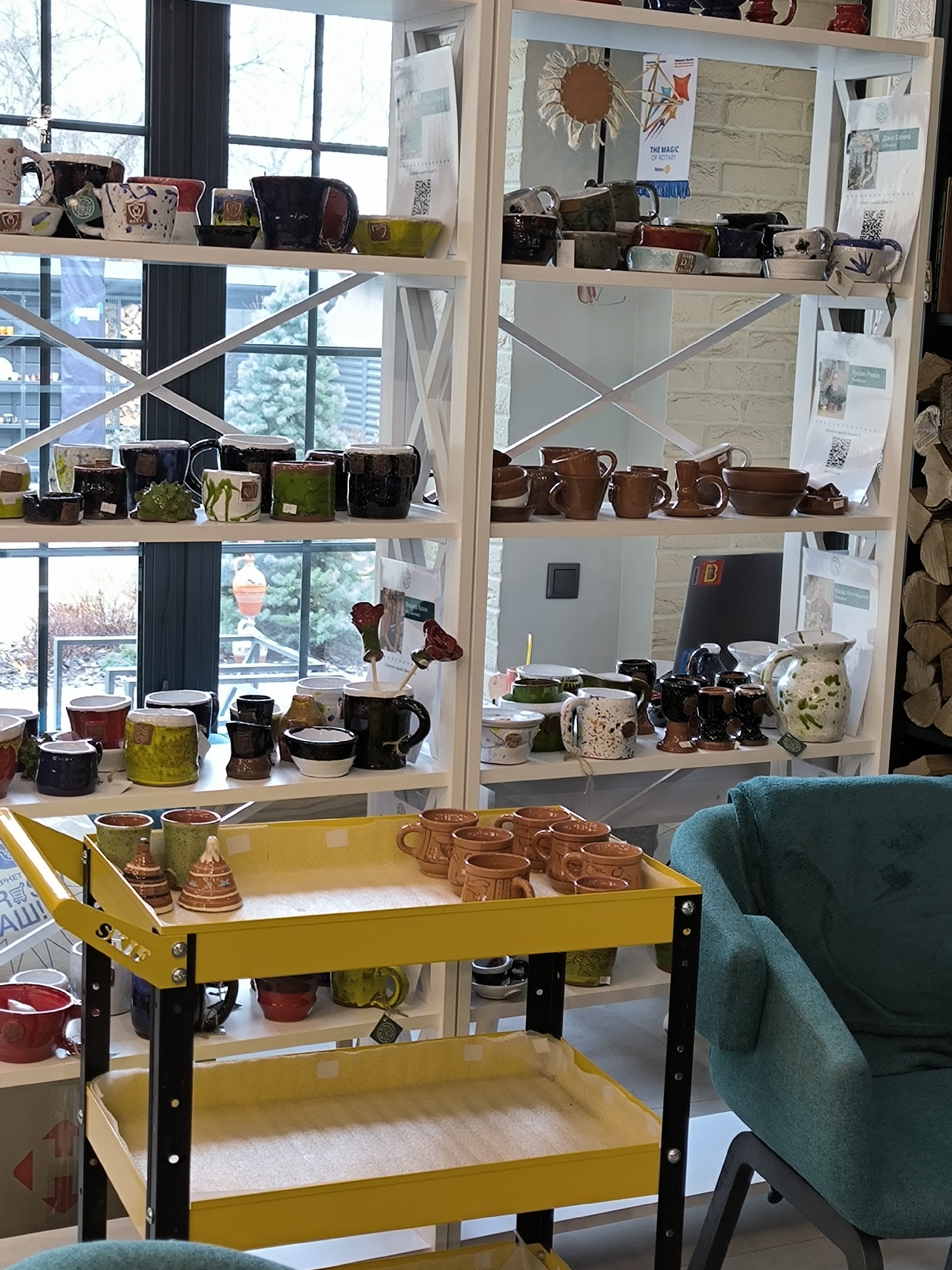 The image shows a display of handmade ceramic items in a shop in Vinnytsia, Ukraine. The items, mostly mugs and bowls in various colors and designs, are arranged on white shelves and a yellow rolling cart with black frame. A green armchair is positioned to the right of the cart. The shelves also hold promotional materials and decorative items. Outside the shop, a street scene is visible through the window.