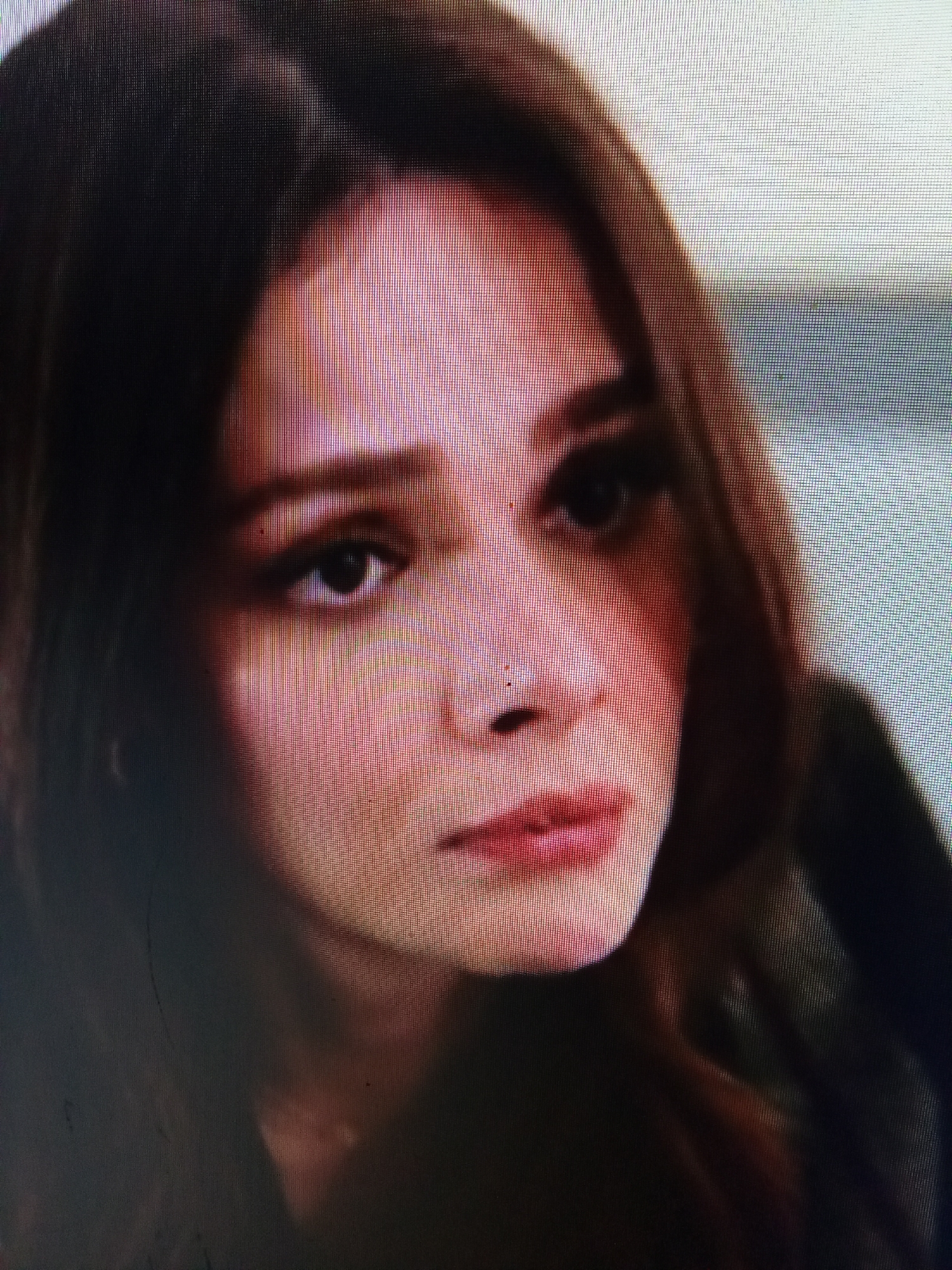 The image is a close-up, low-resolution shot of a young woman's face. She has dark brown hair that is partially obscuring her face, fair skin with a pinkish hue, and dark eyes. Her expression appears to be one of sadness or distress, with her brow furrowed and her mouth slightly open. The lighting is dim, casting shadows on her face and making it difficult to discern fine details. The background is out of focus and appears to be an indoor setting. There is no indication of the location being in Рощин ауылдық округі, Kazakhstan.