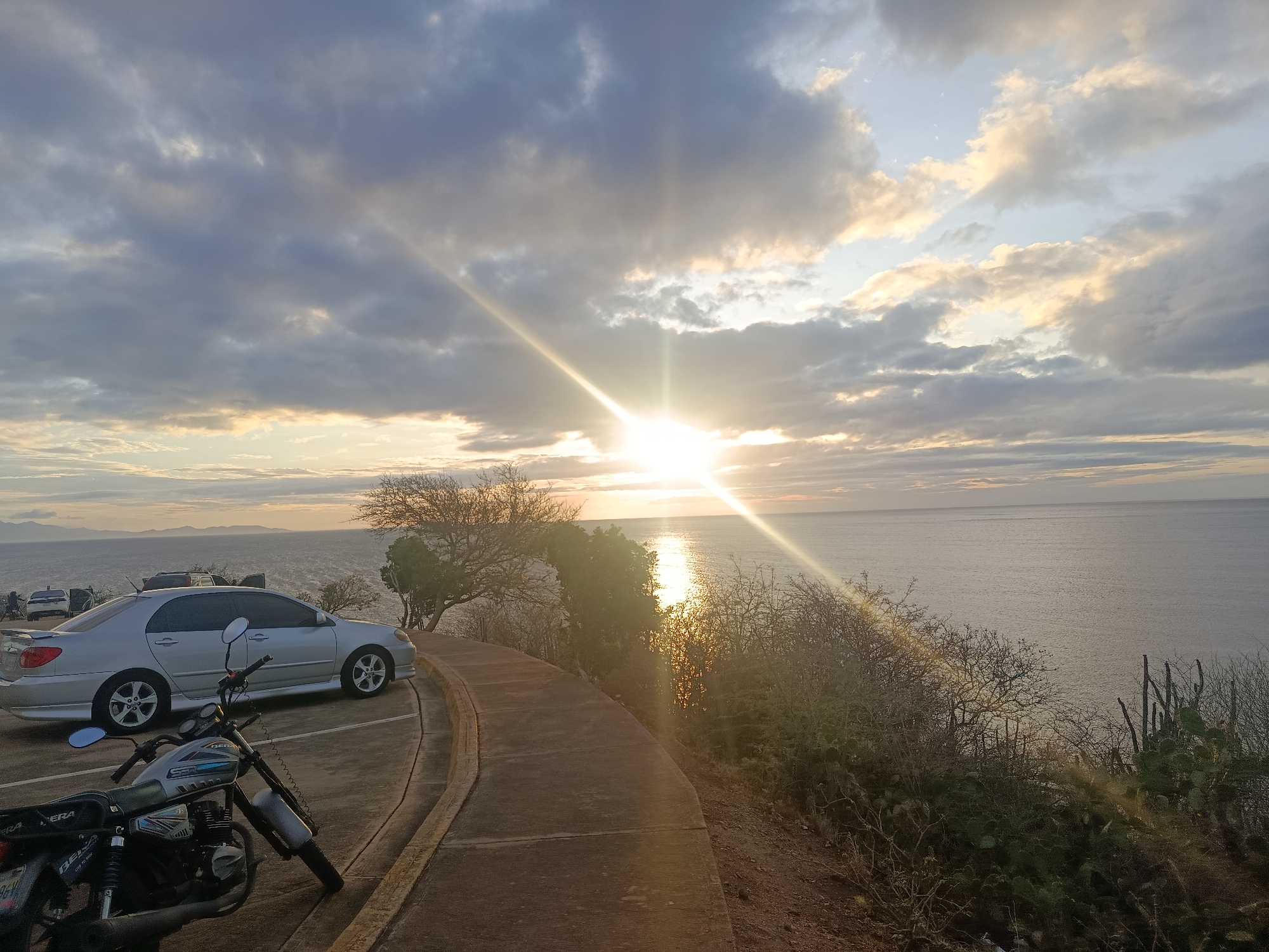 This media file captures a scenic coastal landscape at either sunrise or sunset, viewed from a high vantage point overlooking the ocean, likely in Juan Griego, Venezuela. The setting or rising sun is a prominent feature, positioned centrally in the upper-right portion of the frame, emitting strong golden rays and creating a noticeable lens flare that stretches across the sky and water.
The sky is partly cloudy, displaying a mix of dark gray clouds and clouds illuminated with warm, golden hues by the sun. The vast expanse of the ocean stretches from the mid-ground to the horizon, reflecting the sun's light in a bright, shimmering path across its surface.
In the foreground and mid-ground, a paved path or road curves along the edge of the land. On the left side of this path, a silver sedan, possibly a Toyota Corolla, is parked. Closer to the viewer, in the immediate foreground on the left, a motorcycle is parked, with the brand name "BERA" visible on its side panel. The terrain along the coast is covered with sparse, dry-looking vegetation, including gnarled trees, various bushes, and cacti, indicative of an arid climate.
The overall scene conveys a peaceful moment, emphasizing the natural beauty of the coastline during the golden hour. No people are clearly visible. The strong natural light creates a high contrast between the bright sky and ocean and the more shadowed foreground elements.