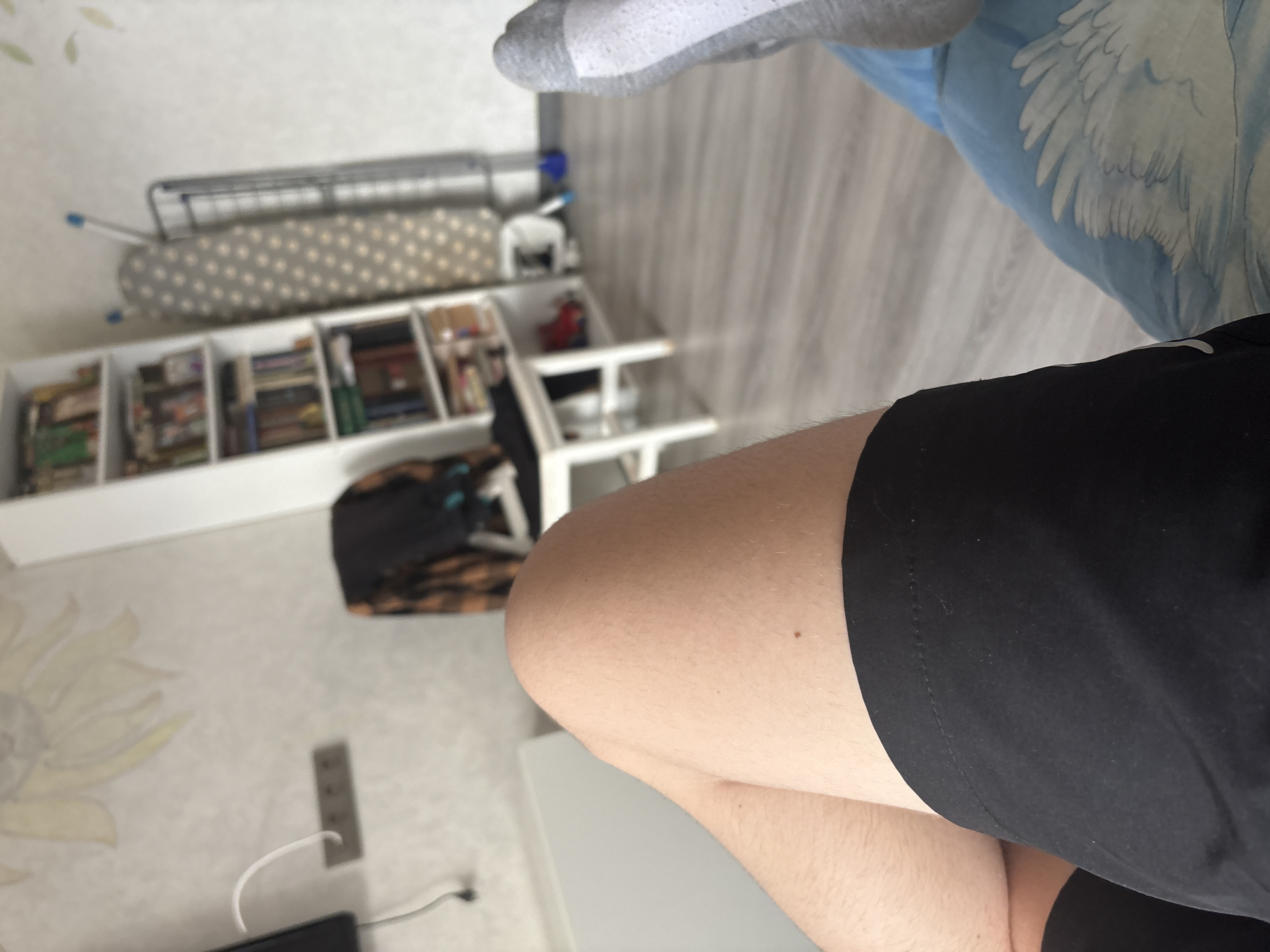 The image shows a close-up view of a person's legs and feet, with a portion of a living room in the background. The person is wearing black shorts and grey and white socks. The background features a bookshelf filled with books, an ironing board, and a chair with clothing draped over it. The flooring appears to be light-colored laminate. The lighting suggests an indoor environment, possibly during the day. The scene is static, with no apparent action or interaction occurring.