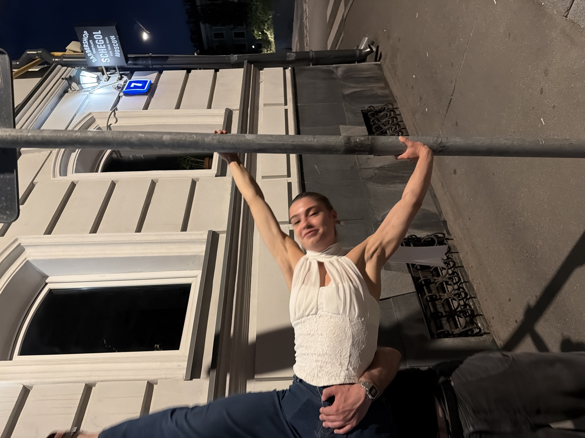 A woman in a white top is being held upside down by a person whose arm is around her waist. She is reaching out and holding onto a metal pole. A sign for "Barbershop Schegol Moscow" is visible on a lamppost to the right of the pole. The building behind them has a light-colored facade with dark windows. The scene appears to be on a street at night.