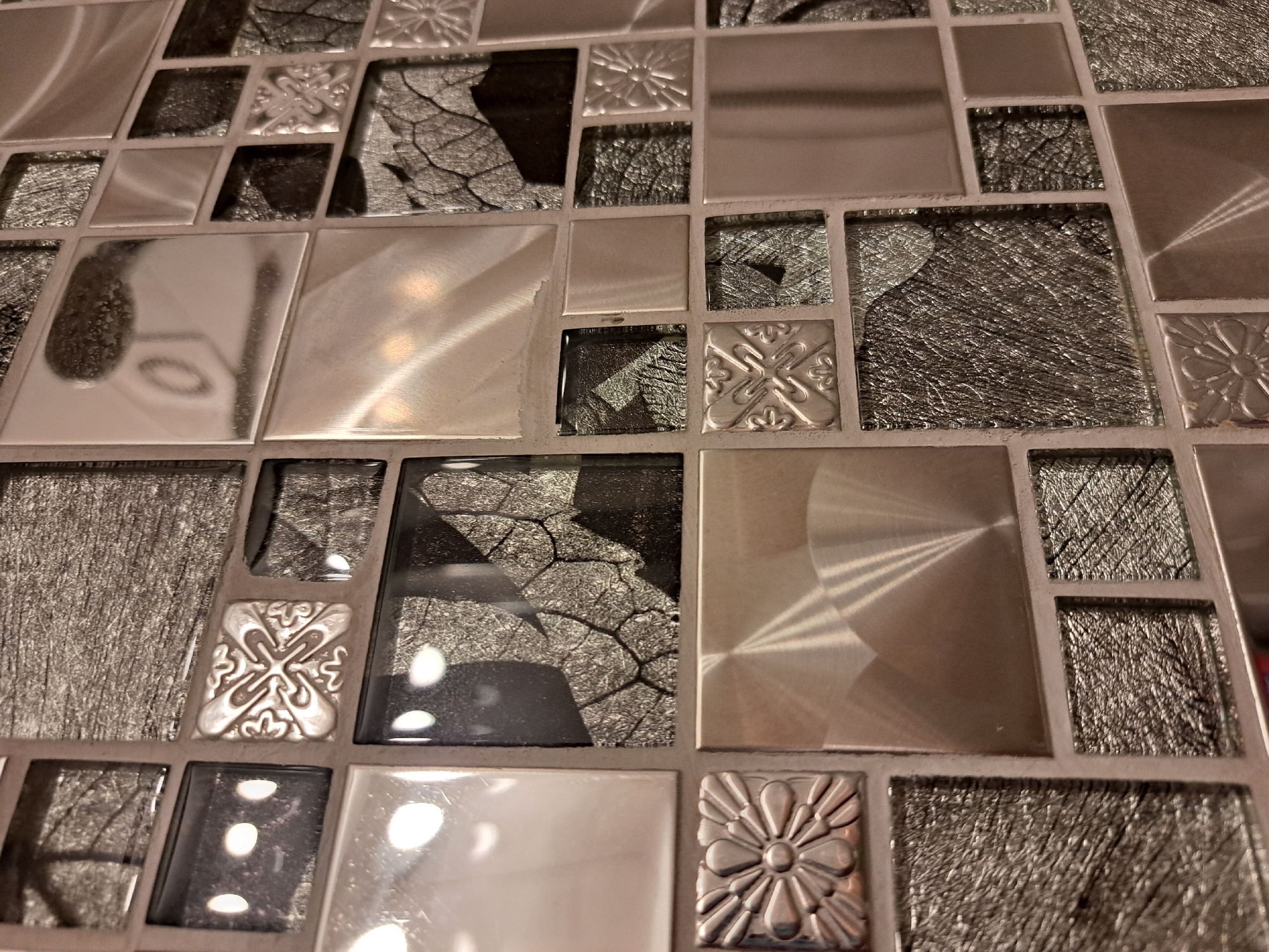 The image displays a close-up, angled view of a tiled wall. The tiles are a mix of finishes and patterns, predominantly in shades of silver, grey, and black. Some tiles are smooth and reflective, showing light reflections that create abstract shapes. Others have a textured, metallic appearance with fine lines or a cracked pattern. There are also a few decorative tiles with embossed floral or geometric designs. The grout lines are visible, forming a grid pattern. The lighting appears to be artificial, casting highlights and shadows that add depth and texture to the surface. There is no indication of people, actions, or interactions. The image is a still shot of a decorative tile installation. There is no information in the image to suggest a location in Irkutsk, Russia.