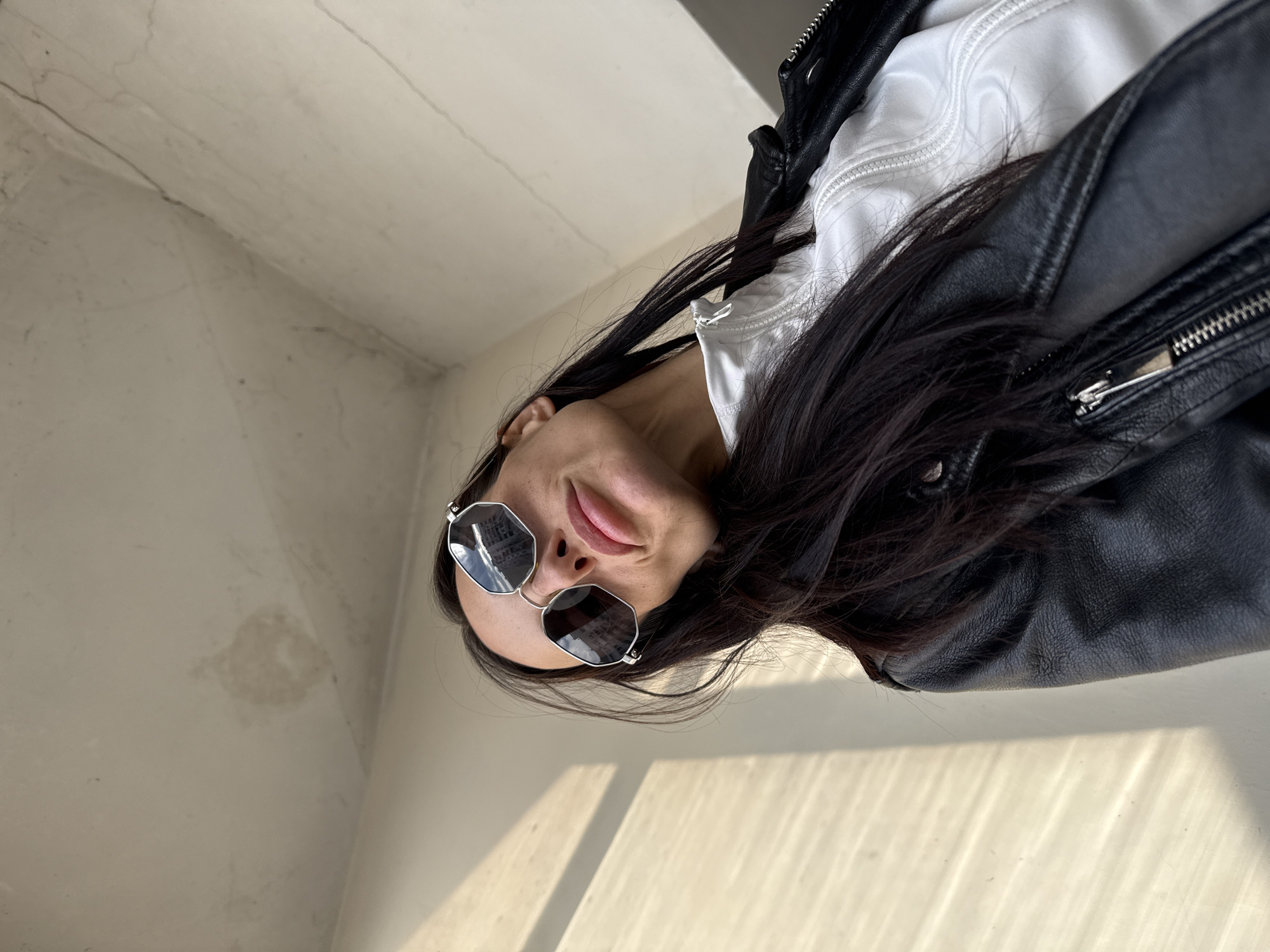 This image, set in Pleven, Bulgaria, presents an adult female photographed from a low angle against a corner formed by two light-toned walls. The subject has long, dark hair and wears octagonal dark-lensed sunglasses with silver-colored frames; these lenses reflect building facades. She wears a black leather jacket layered over a white garment featuring a zipper. Her lips are light pink. The walls exhibit surface wear, including fine cracks, minor discoloration, and a prominent circular water stain above the subject's head. Direct sunlight casts distinct horizontal and vertical shadows on the wall to the left of the subject.