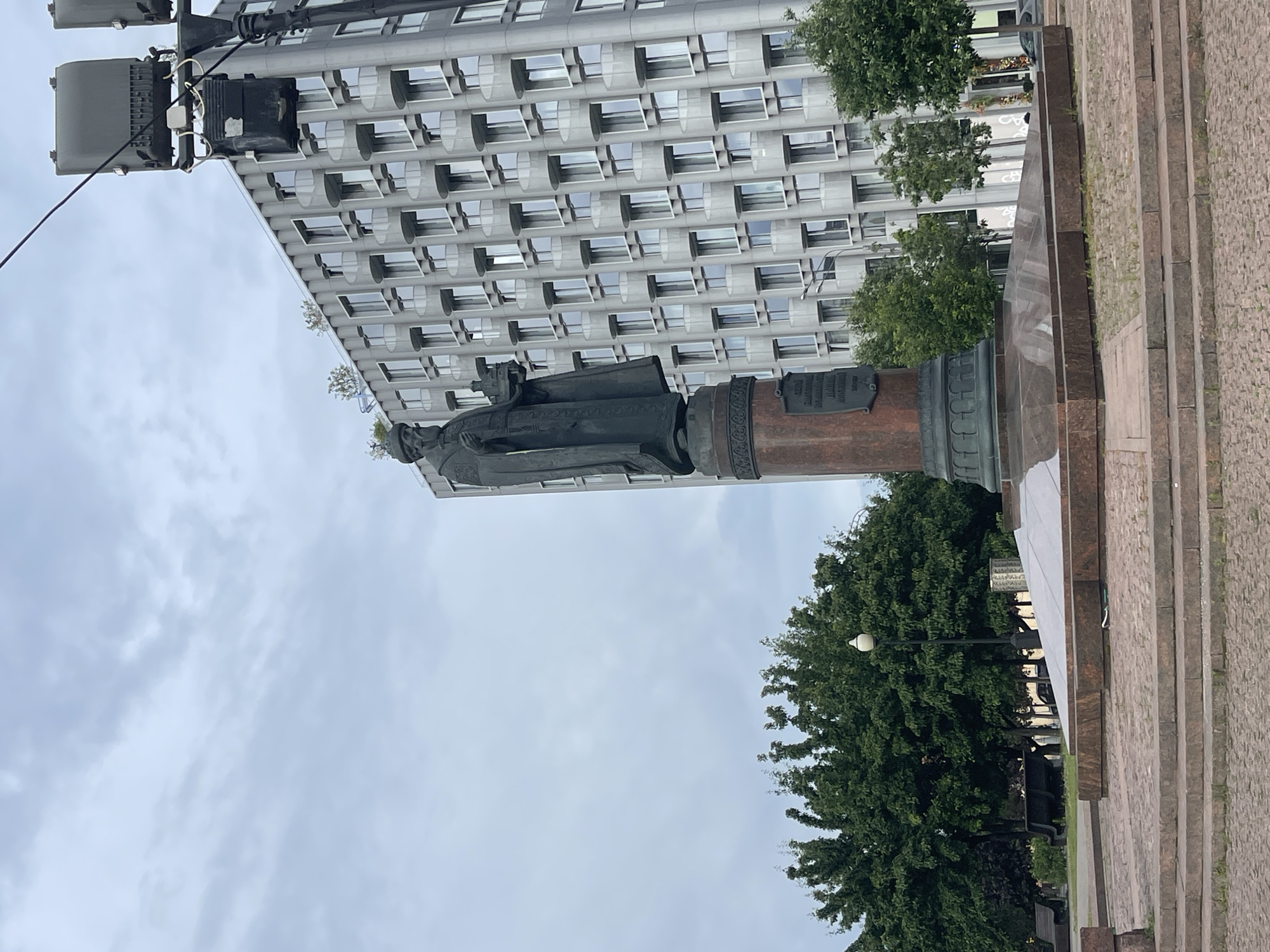 The image depicts the monument to Ivan Fedorov in Moscow, Russia. A bronze statue of Fedorov stands on a reddish-brown pedestal, with a plaque visible on the pedestal's side. Behind the monument is a modern building with a gridded facade. Trees are situated around the monument, and a cloudy sky is visible. A lamppost and steps are present near the monument's base. A street lamp is visible in the top-left.