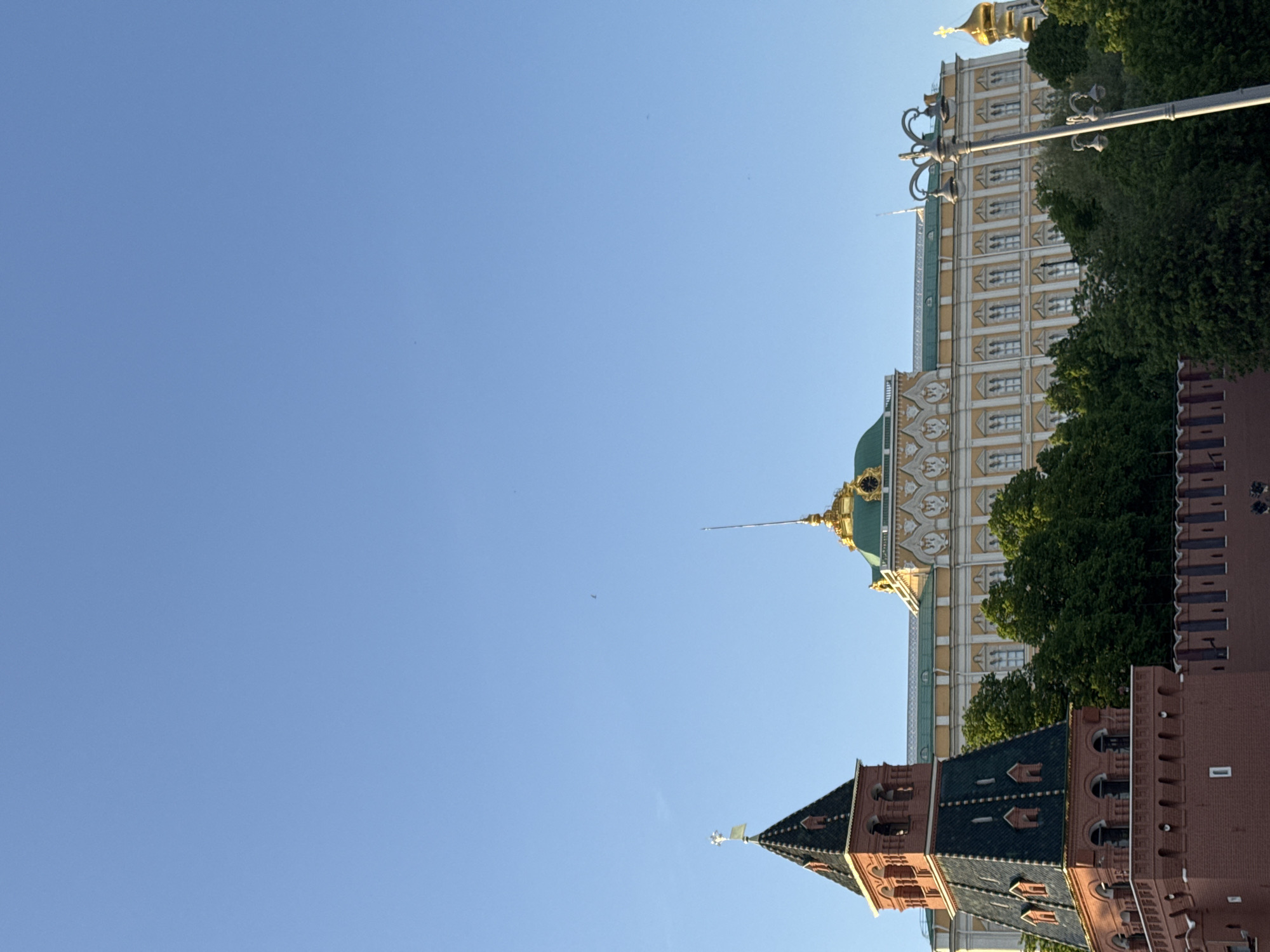 The image shows a view of the Moscow Kremlin in Moscow, Russia, with the blue sky occupying most of the frame. The architecture includes the Ivan the Great Bell Tower, the Grand Kremlin Palace, and portions of the Kremlin walls. Trees are interspersed throughout the scene.