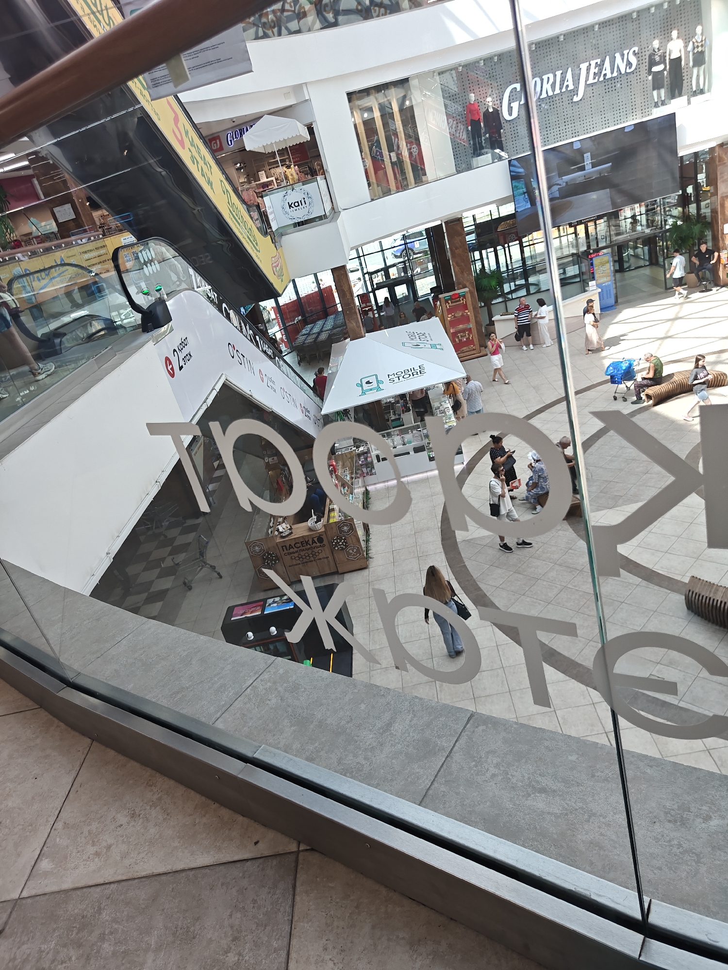 The image shows an interior view of a shopping mall in Petropavl, Kazakhstan. The perspective is from an elevated position, looking down through a glass partition with white lettering on it.
In the background, an escalator is visible, with yellow signage on its side. Various retail stores are present, including "Gloria Jeans" with mannequins displayed in the window. Other stores are partially visible, some with umbrellas or awnings. People are seen walking around the mall, some are sitting on benches, and others are standing in groups. A blue shopping cart is also present.
The foreground is dominated by the glass partition with large white stylized lettering that appears to be part of the mall's branding. Some of the letters are obscured by the reflection and the angle of the shot, but the general shape suggests words or logos. The floor of the mall is tiled.
The overall impression is a busy commercial environment.