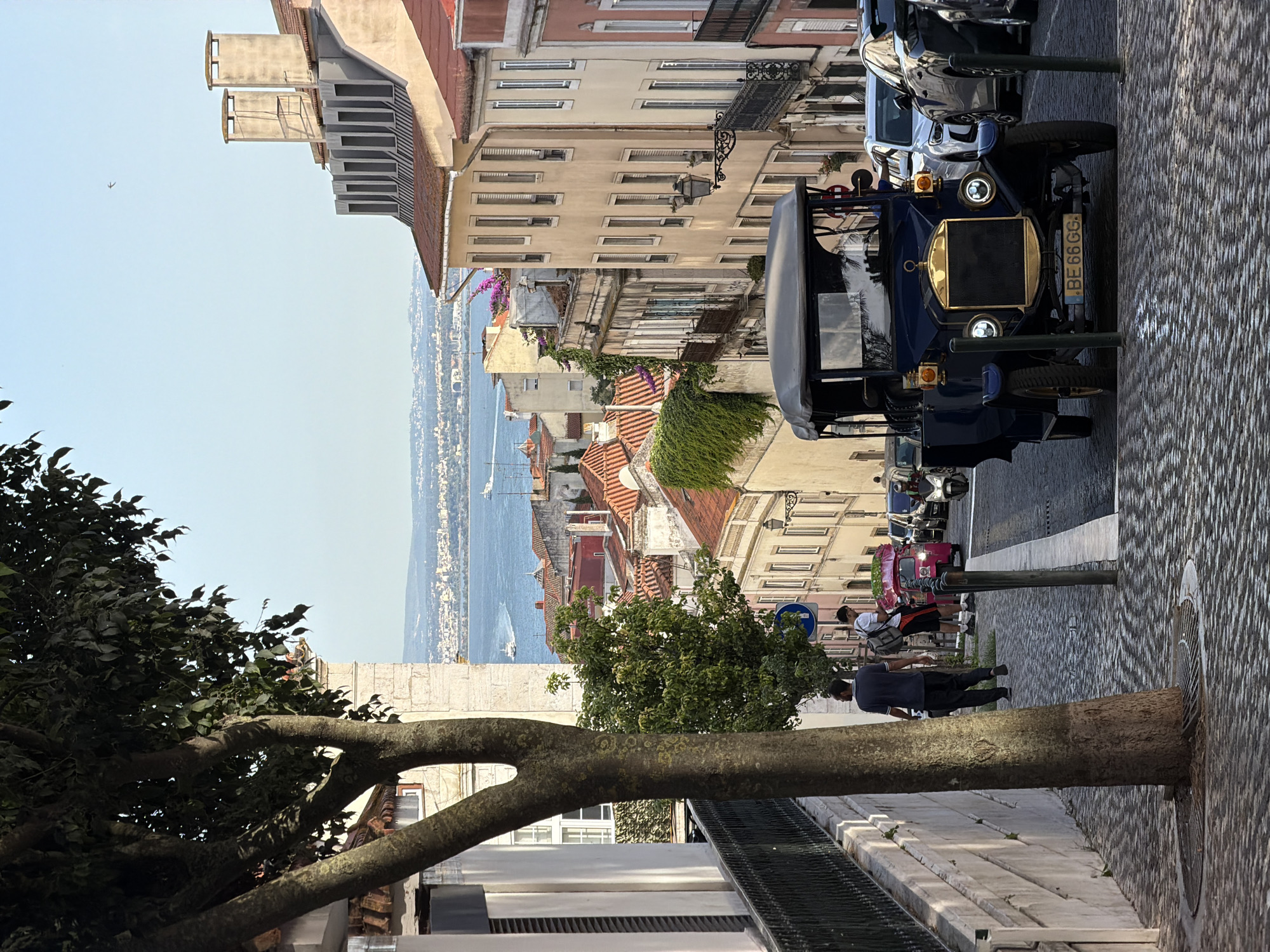 The image depicts a street scene in Lisbon, Portugal, looking down a cobblestone slope towards the Tagus River and the city skyline. In the foreground, a vintage dark blue car with a canvas top is parked, its license plate reading "BE 66 GG". Behind it, modern cars and a pink tuk-tuk are also visible. Two men are walking up the street on the left side, one in a dark shirt and pants, the other in a white t-shirt and shorts with a backpack. A large tree trunk with dark bark is prominent on the left, with its branches extending into the upper left corner of the frame. The buildings lining the street are typical of Lisbon, with colorful facades, red-tiled roofs, and intricate wrought-iron balconies. The view opens up to the Tagus River, with boats sailing on its waters, and the distant cityscape of Lisbon under a clear blue sky. A single bird is flying in the sky above the buildings. The atmosphere appears calm and sunny.