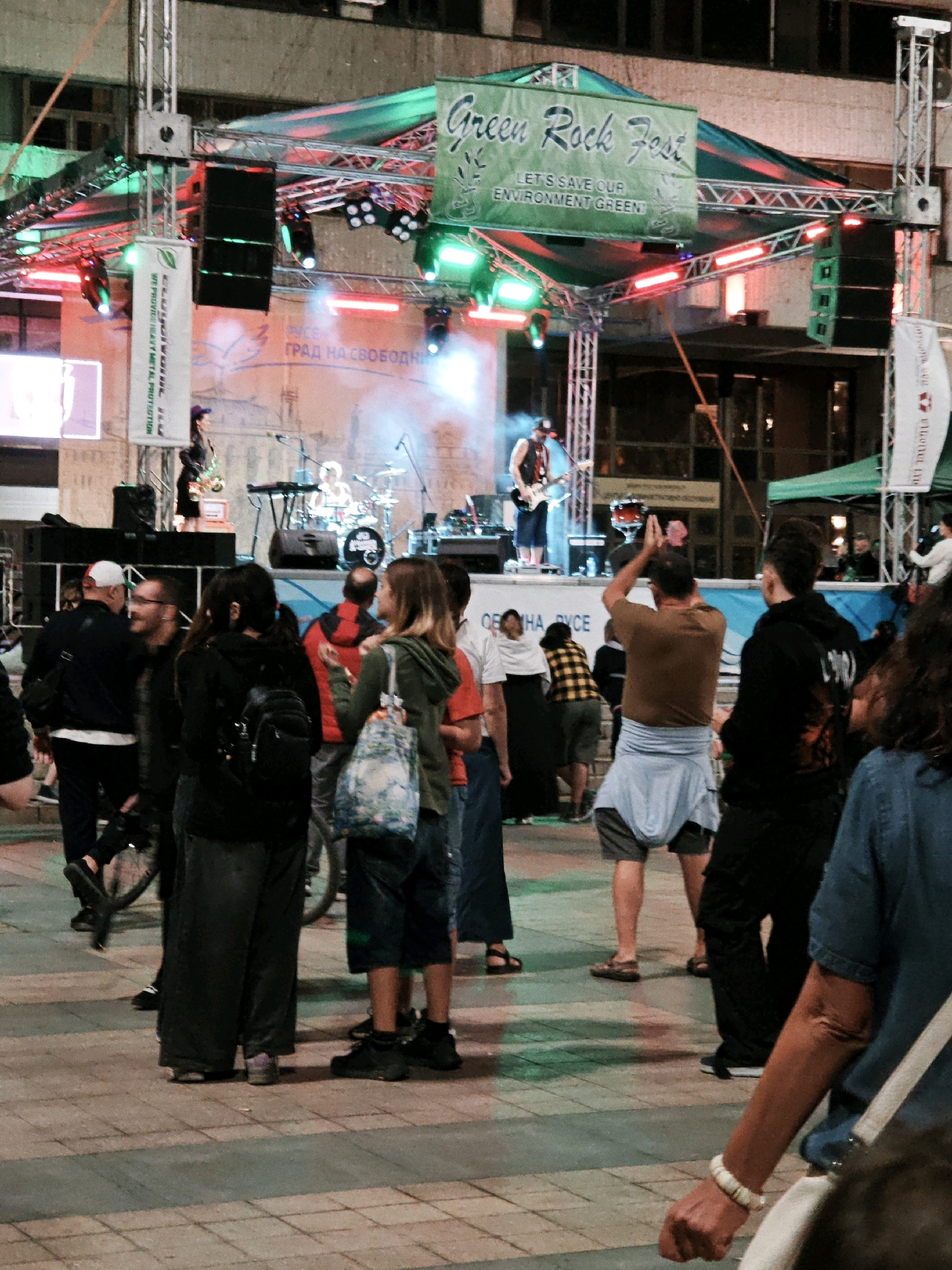 The image captures an outdoor music festival event, likely in a city square or plaza given the paved ground and surrounding buildings. A stage is set up with a banner that reads "Green Rock Fest" and the tagline "LET'S SAVE OUR ENVIRONMENT GREEN." Musical equipment, including speakers and instruments, are visible on the stage, and a band is performing. A crowd of people is gathered in front of the stage, some watching the performance, while others are mingling. Various individuals in the crowd are dressed in casual attire. The lighting appears to be a mix of stage lights and ambient evening illumination. The environment suggests a public gathering space, possibly at night or during late afternoon, with the architecture of the background buildings indicating an urban setting.