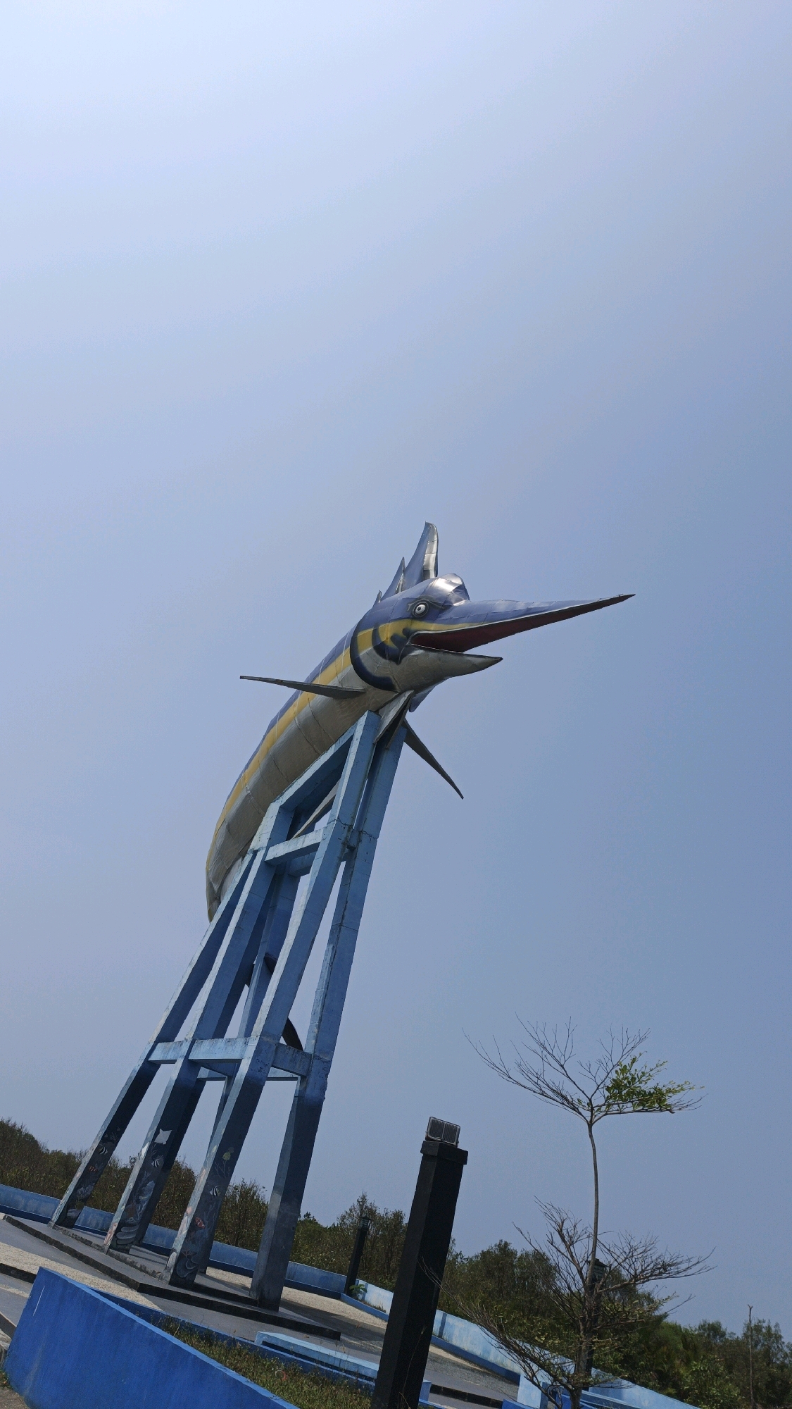 This is a medium-angle, vertical outdoor shot of a large marlin sculpture on a blue metal support structure, set against a clear, pale blue sky. The sculpture depicts a marlin with metallic gray scales, yellow stripes, and blue fins, its mouth open and bill pointed to the right. The blue support structure is triangular and made of thick beams. Below the sculpture, there are blue concrete barriers, paved walkways, and patches of greenery. In the background, there is a line of trees, and a small, bare tree with a few green leaves is visible to the right of the support structure. The scene appears to be a public monument or park in Pangandaran, Indonesia, likely near the coast, given the prominent marlin sculpture which is a symbol of the area. There are no people or animals visible in the image.