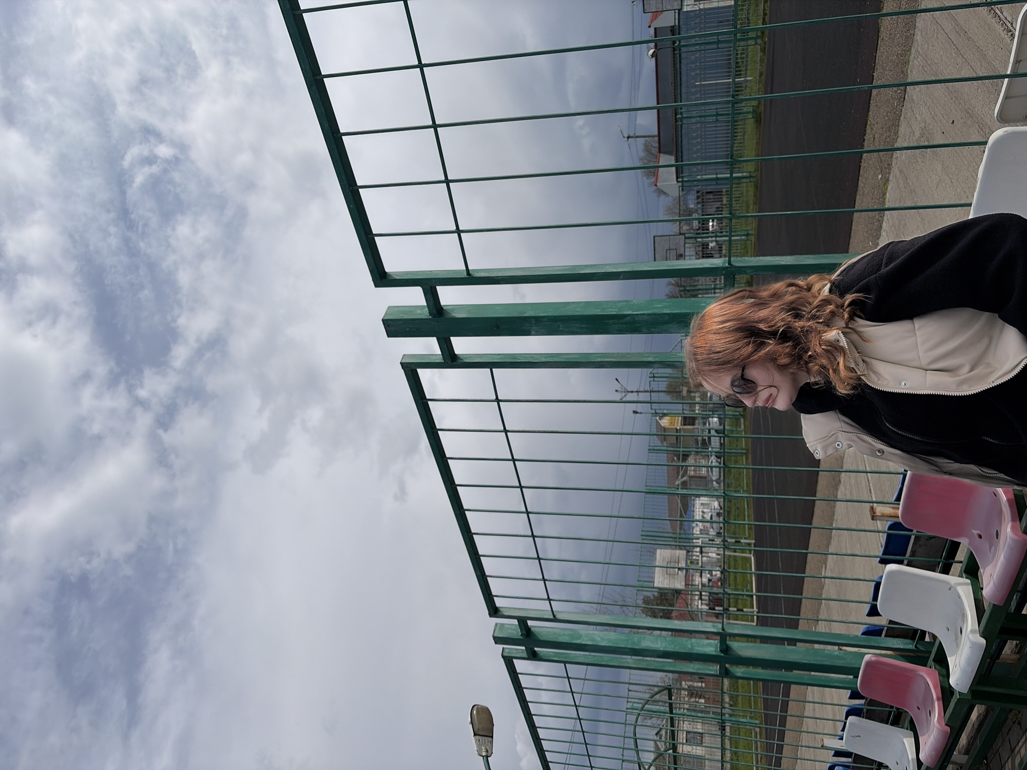 A low-angle shot shows a woman with red curly hair, wearing sunglasses, a black jacket, and a beige vest, seated in a stadium. The seating arrangement includes alternating pink, white, and blue plastic seats. A green metal fence frames the view. In the background, a city landscape unfolds under a cloudy sky. Utility poles and various buildings are visible. The location is Novopokrovskaya, Russia.