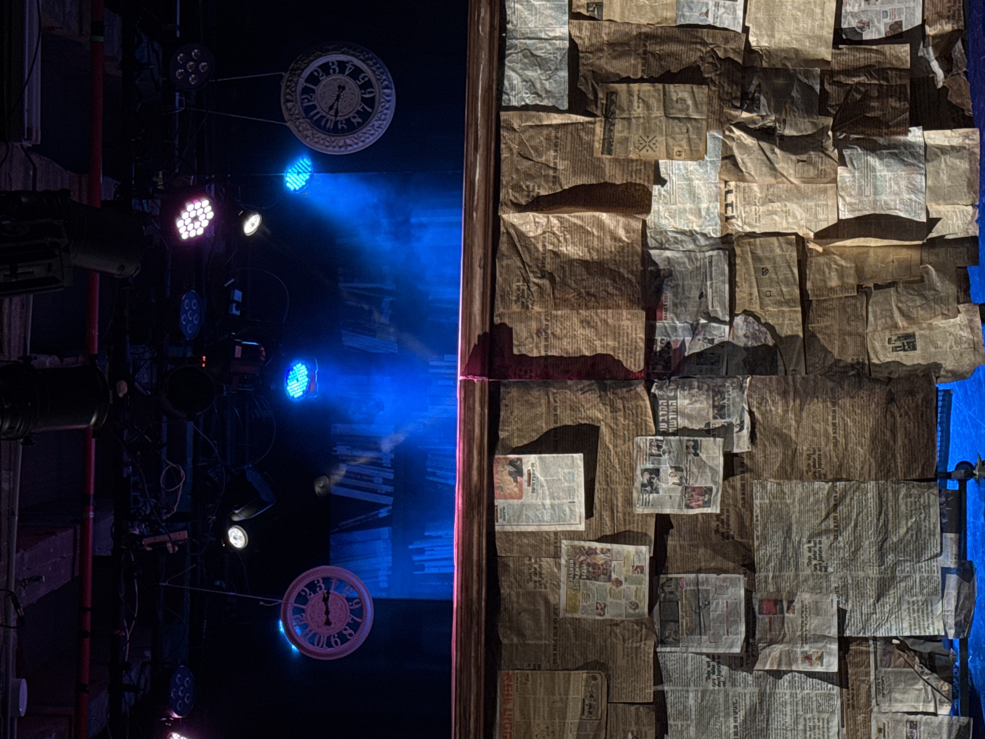 The image shows a stage set featuring a dark, industrial left side with exposed lighting equipment, two decorative clocks, and blue lighting effects. The right side is covered in crumpled newspaper pages, some of which have images and text in Hebrew. The set appears to be for a theatrical production. The image does not provide any information related to a location in Tel-Aviv, Israel, aside from the possibility of the text in the newspapers being Hebrew.