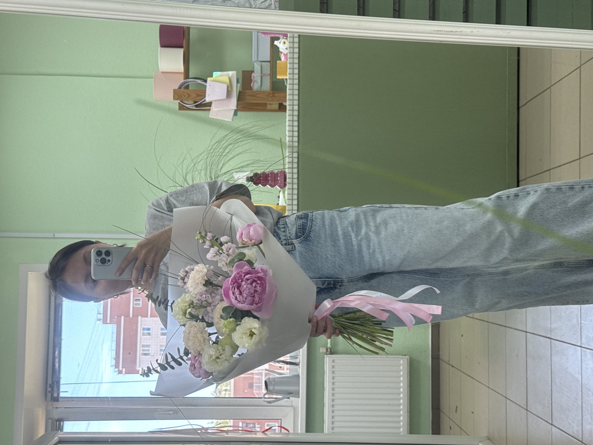 A woman holds a bouquet of flowers while taking a selfie in front of a mirror. The reflection in the mirror shows a building in the city of Izhevsk, Russia. The bouquet consists of pink and white flowers wrapped in white paper and tied with a pink ribbon. The woman is wearing a gray shirt and blue jeans. The background includes a green wall, a window, and a white radiator.