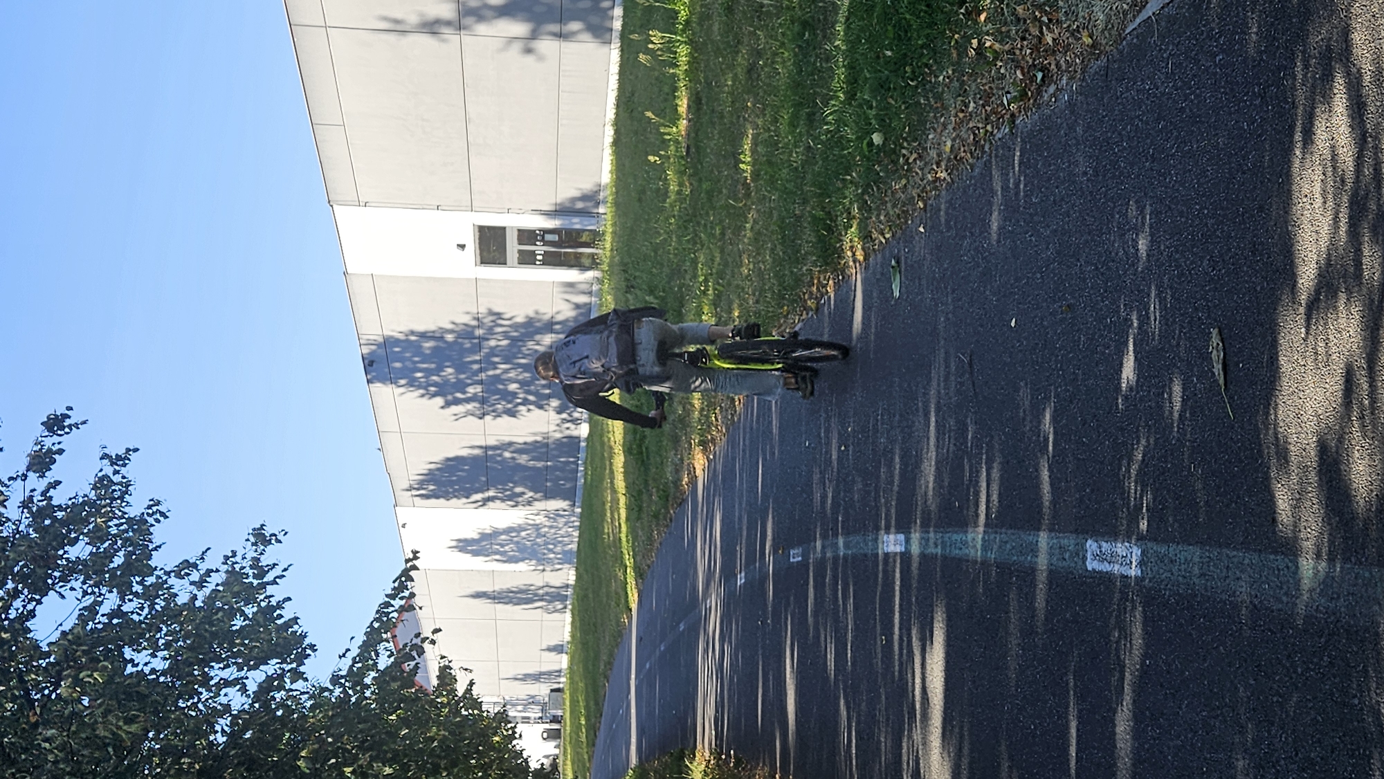 A person is riding a bicycle on a paved bike path, viewed from behind. The cyclist wears a backpack and light-colored pants. The bike has a fluorescent yellow frame. The path is marked with white and green lines. To the right of the path is a grassy area with some fallen leaves. Behind the grass is a long, light-colored building with a modern facade, featuring a segmented design and a few windows or doorways. Shadows of trees are cast on the building's exterior. To the left of the path, a large tree with dark green leaves extends into the frame. The sky is clear and blue. This scene is located on the bike path adjacent to the National Stadium (Štadión Toryská) in Prešov, Slovakia.