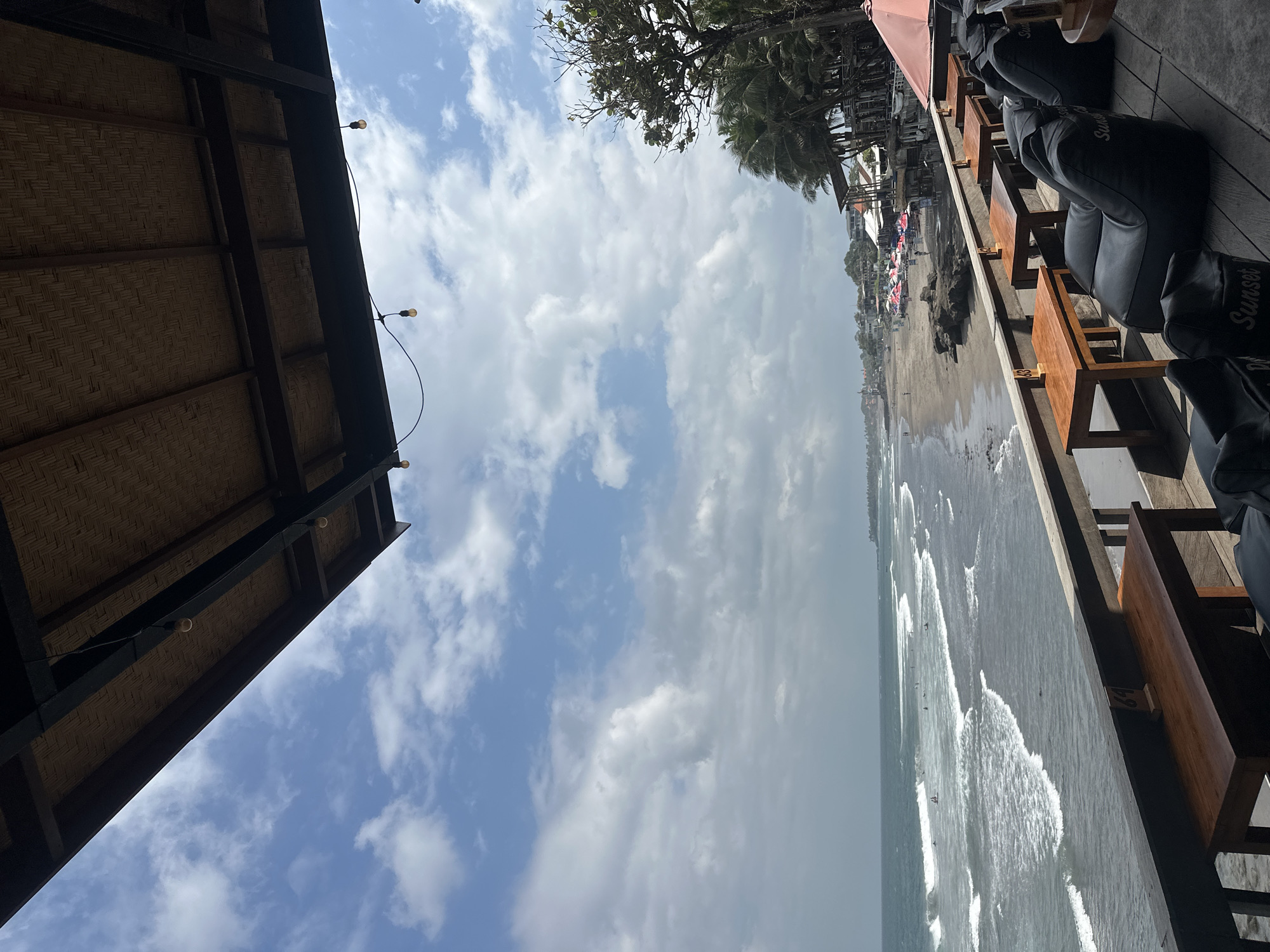 The image captures a view from a rooftop cafe overlooking a beach and ocean. In the foreground, several black beanbag chairs and small wooden tables are arranged on a wooden deck. A brown wooden table with a number "63" sign is visible. The cafe is covered by a thatched roof supported by dark metal beams, with exposed light bulbs hanging from the ceiling.
Beyond the cafe, a sandy beach stretches along the coastline, dotted with beach umbrellas and a few people. The ocean is visible with moderate waves breaking on the shore. Several surfers are in the water. Palm trees and other vegetation line the beach, behind which buildings and structures of a resort or village are apparent. The sky is partly cloudy with a mix of blue sky and white clouds. The scene depicts a casual, tropical beachside setting in North Kuta, Indonesia, likely a popular spot for beachgoers and cafe patrons.