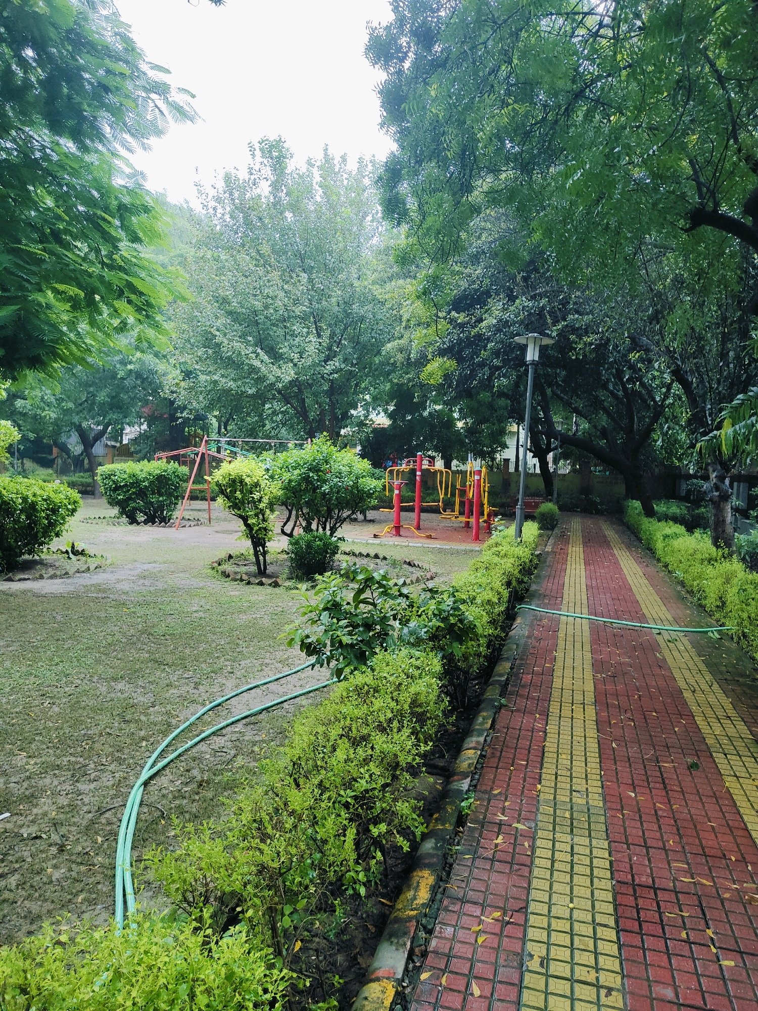 The image depicts a park scene in Gurgaon, India. A paved walkway, made of red and yellow tiles, runs diagonally from the bottom right towards the center of the frame. Lush green bushes line the left side of the walkway. On the left side of the walkway, a grassy area with scattered trees and bushes contains playground equipment, including yellow and red exercise machines and a swing set. Two green hoses lie coiled on the wet grass. The sky is overcast, suggesting a cloudy or rainy day. The overall impression is that of a well-maintained public park.
