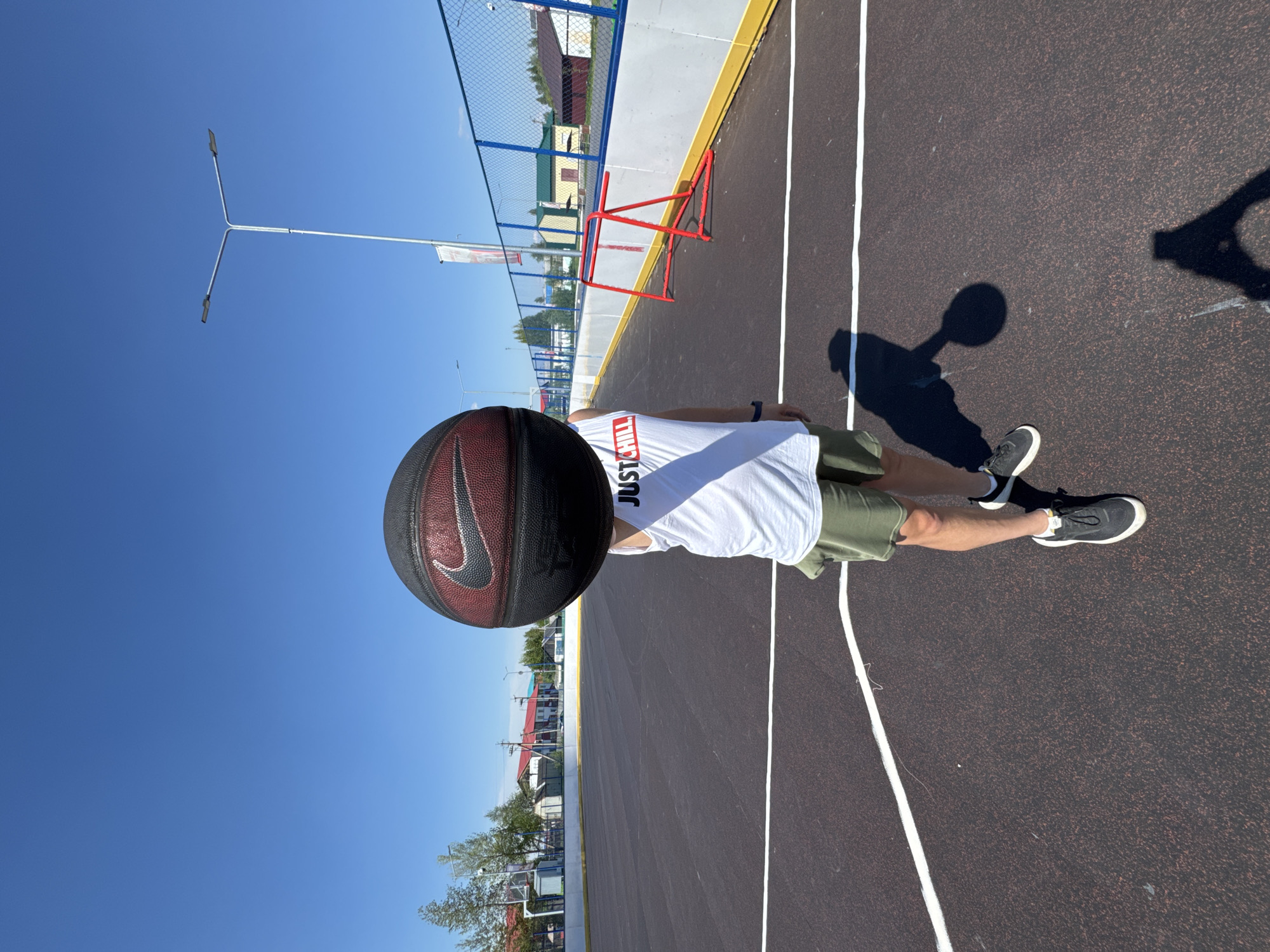 This image, captured in Высокий, Russia, depicts a clear, sunny day on an outdoor basketball court. A person, visible from the chest down, stands on the dark-surfaced court marked with white lines. They are wearing a white t-shirt with the red text "JUST CHILL," olive green shorts, and black athletic shoes. The person is holding a black and reddish-brown basketball featuring a prominent black Nike swoosh logo.
The bright sunlight casts distinct shadows on the court, including the person's shadow to their left and a larger shadow resembling a basketball hoop structure further to the right. In the background, under a cloudless blue sky, a silver Y-shaped street light pole with two lamps stands on the left. A blue-framed wire mesh fence separates the court from what appears to be a residential or town area, with various buildings and some trees visible. A small, red metal frame, possibly a goal or part of the court equipment, is also seen near the fence. The scene portrays a casual recreational setting at a community sports facility during daytime.