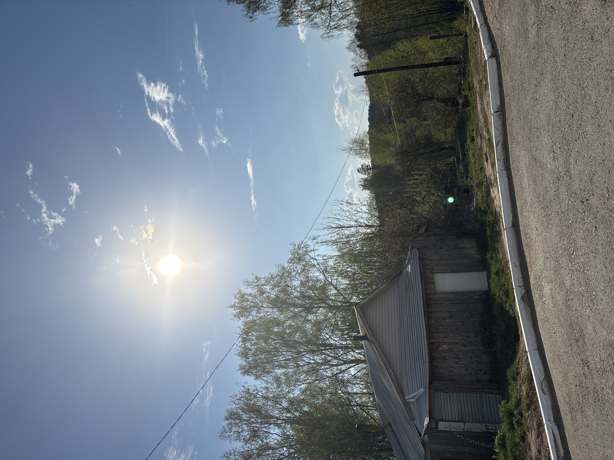 The image presents a view of Chaikovsky, Russia, featuring a landscape with an old wooden building adjacent to an asphalt road. The sky is blue with scattered white clouds and the sun is prominently visible, creating a bright glare. Trees and other vegetation line the background. There are no people visible. A utility pole and an overhead power line are present.