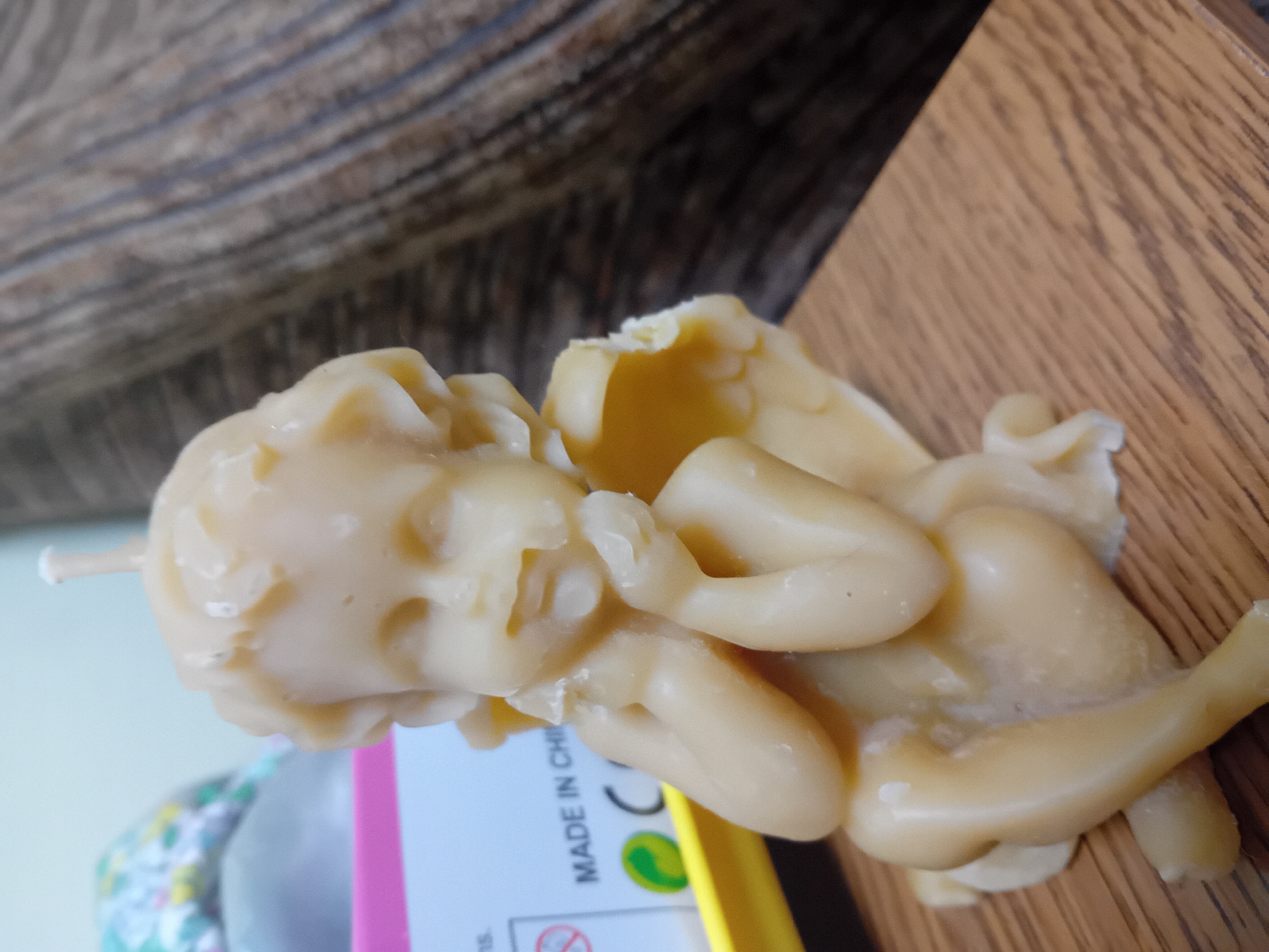 A close-up, side-view, eye-level shot captures a light yellow, cherubic-shaped beeswax candle in focus. The candle is sculpted to depict a winged infant with its hands near its face in a thoughtful or sleeping pose, and it is seated on a wooden surface. In the background, a pink and blue box with some text, including "MADE IN CHI," is visible, as well as a dark, textured surface that could be furniture. The lighting is soft, highlighting the details of the candle's wax. The image is a still life, likely taken indoors, and does not contain any elements related to the city of Chuhuiv, Ukraine.