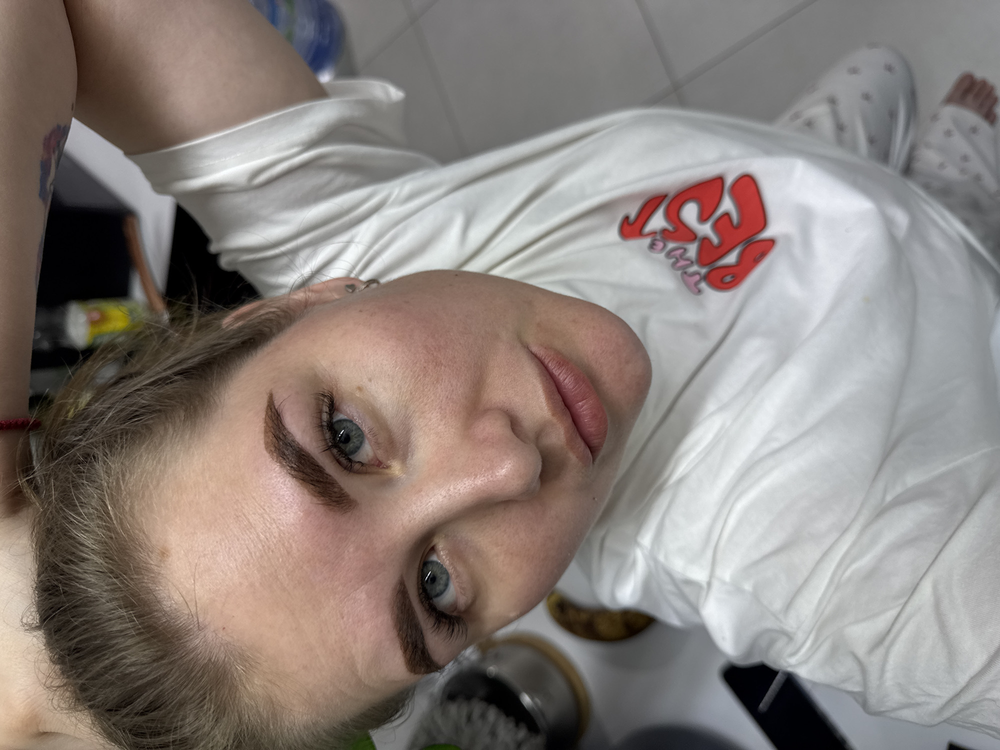 A person is lying down, facing the camera. They have blue eyes and brown eyebrows. The person is wearing a white t-shirt with a red and pink design on it. The person has patterned pajama pants. The location is an indoor setting in Gornyi (Dvurechenskoe S/P), Russia.