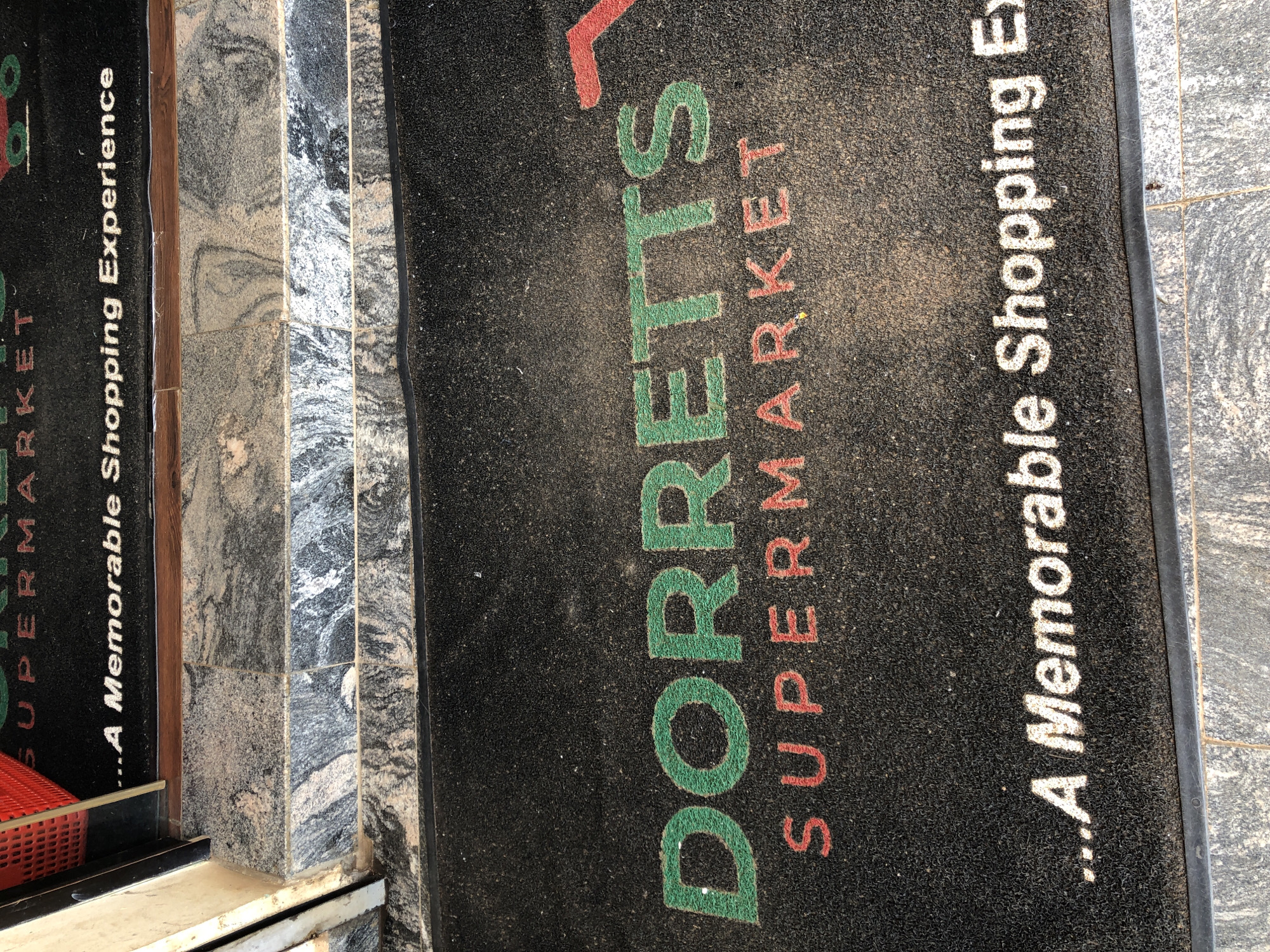 The image shows a close-up of a black welcome mat outside a supermarket. The mat has white text that reads "...A Memorable Shopping Experience". Below this, in larger green letters, it says "DORRETTS" and in red letters, "SUPERMARKET". The supermarket appears to be located in Bar Jirgi Summa, Nigeria, based on previous context. The mat is slightly dusty, and the entrance to the store is visible in the background, with granite-like steps leading up to it.
