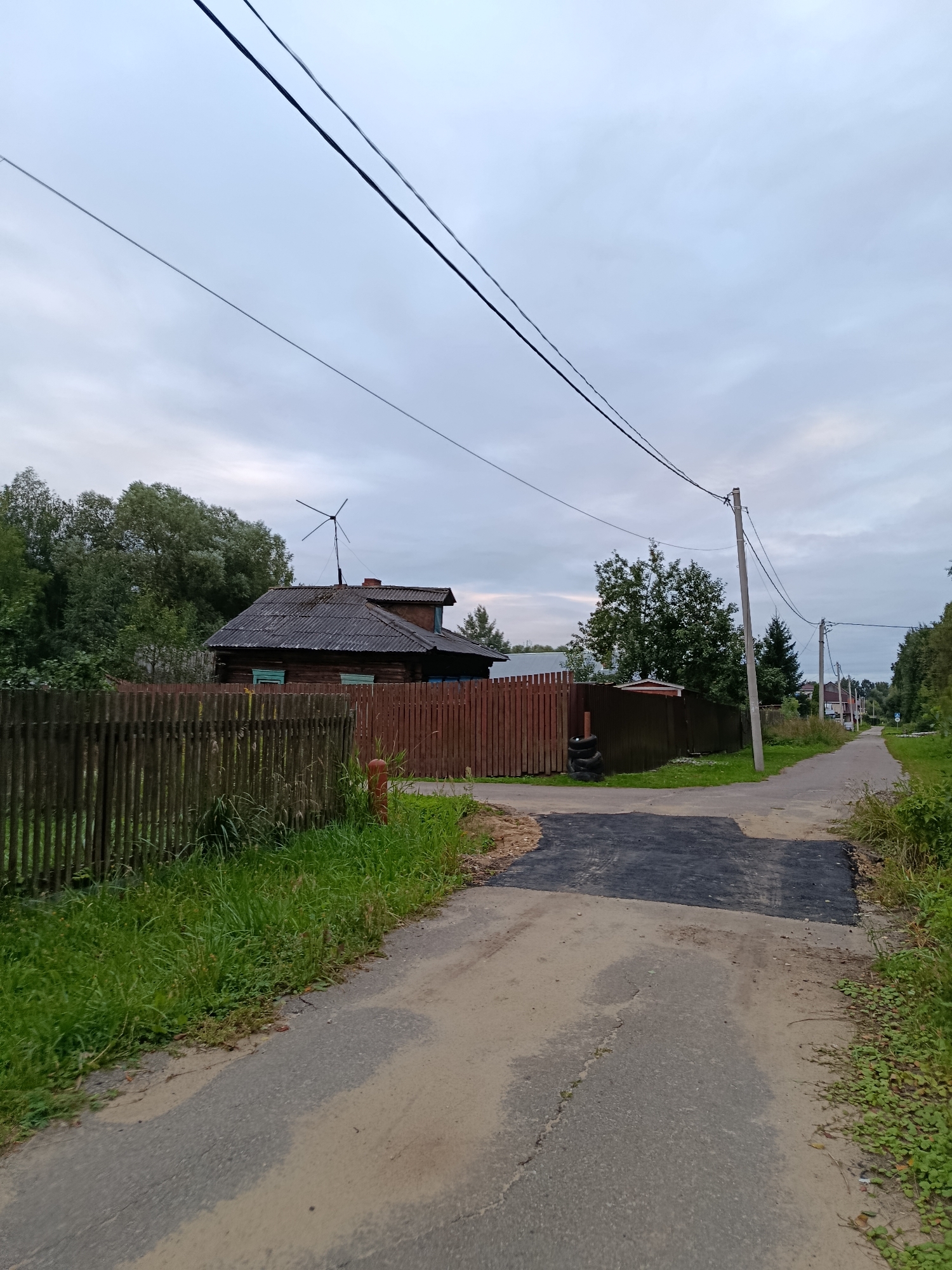 The image depicts a rural street scene in the city of Vereya, Russia, under an overcast sky. In the foreground, a dirt and asphalt road diverges. To the left, a wooden fence borders a property with a weathered wooden house and dense green trees behind it. A small wind turbine is visible on the roof of the house. To the right of the road, a utility pole supports overhead wires, and the road continues into the distance, lined with more trees and vegetation. The overall impression is one of a quiet, undeveloped area.