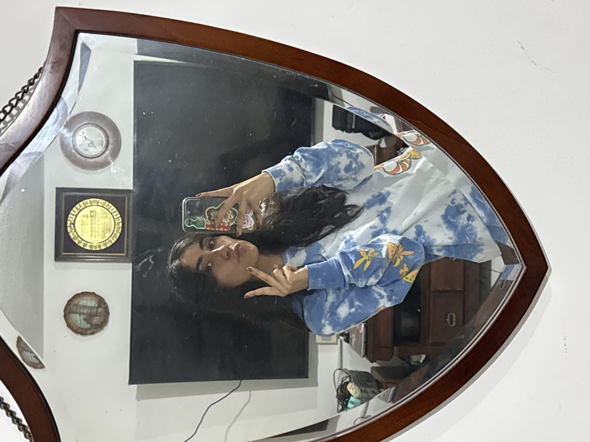 A person is taking a selfie in a mirror. The person has long dark hair and is wearing a blue and white tie-dye sweatshirt with a design on the sleeve. They are holding a phone with a decorated case. The background reflected in the mirror shows a wall with several objects mounted on it, including a round clock or decoration, a framed certificate, and a dark rectangular object that might be a television screen. There's also a glimpse of furniture behind the person. The mirror itself has a dark wooden frame in an unusual shape. The location is not evident in this image.