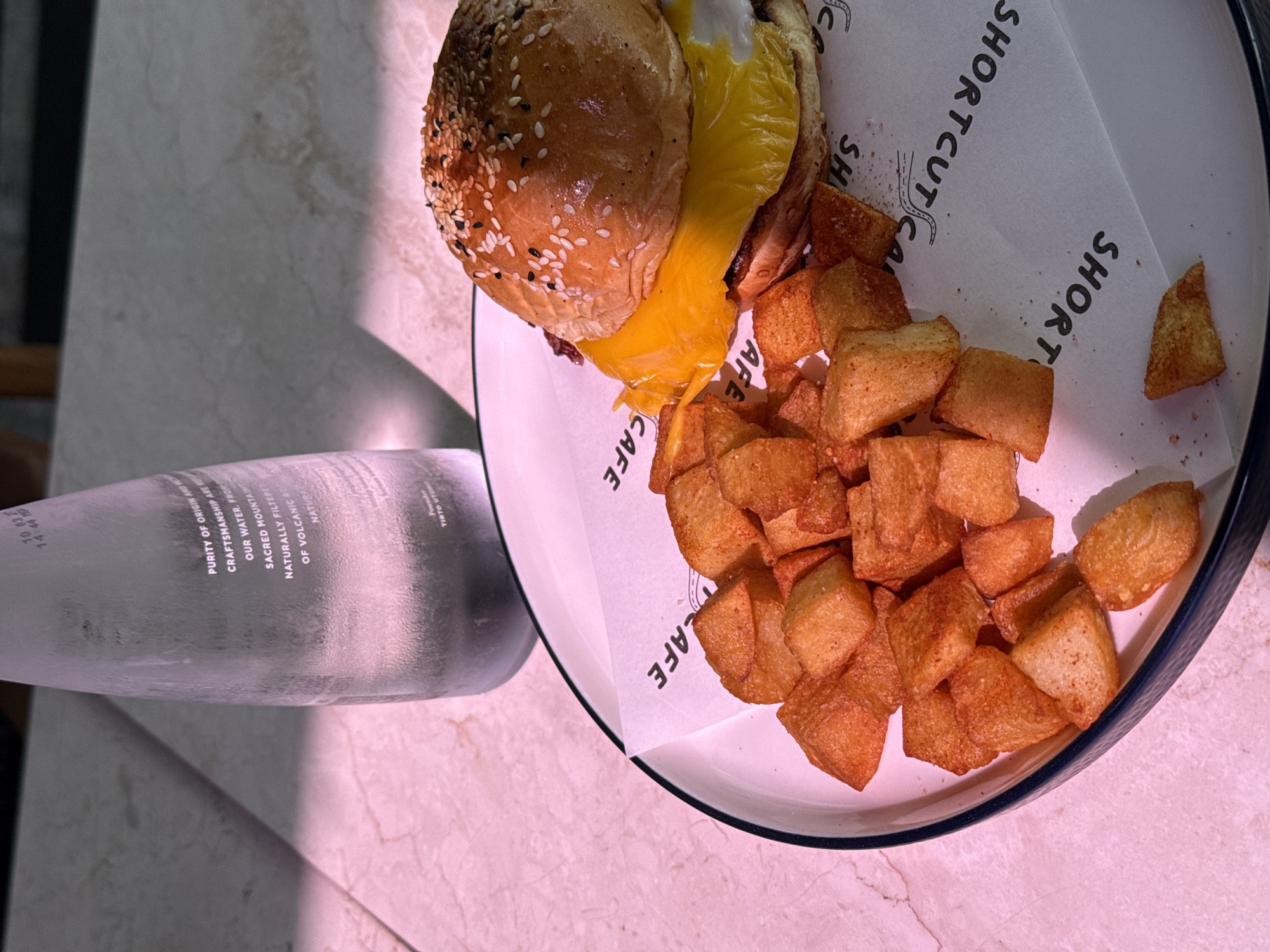 A plate presents a burger with melted cheese and a side of cubed, golden-brown potatoes. The plate is lined with paper printed with "SHORTCUT CAFE," suggesting the meal is from this establishment. A bottle of water rests on the table next to the plate. The water bottle label indicates it's from "TIRTO UTOMO." The location, based on the cafe's name, is likely Canggu, Indonesia, where Shortcut Cafe is located.