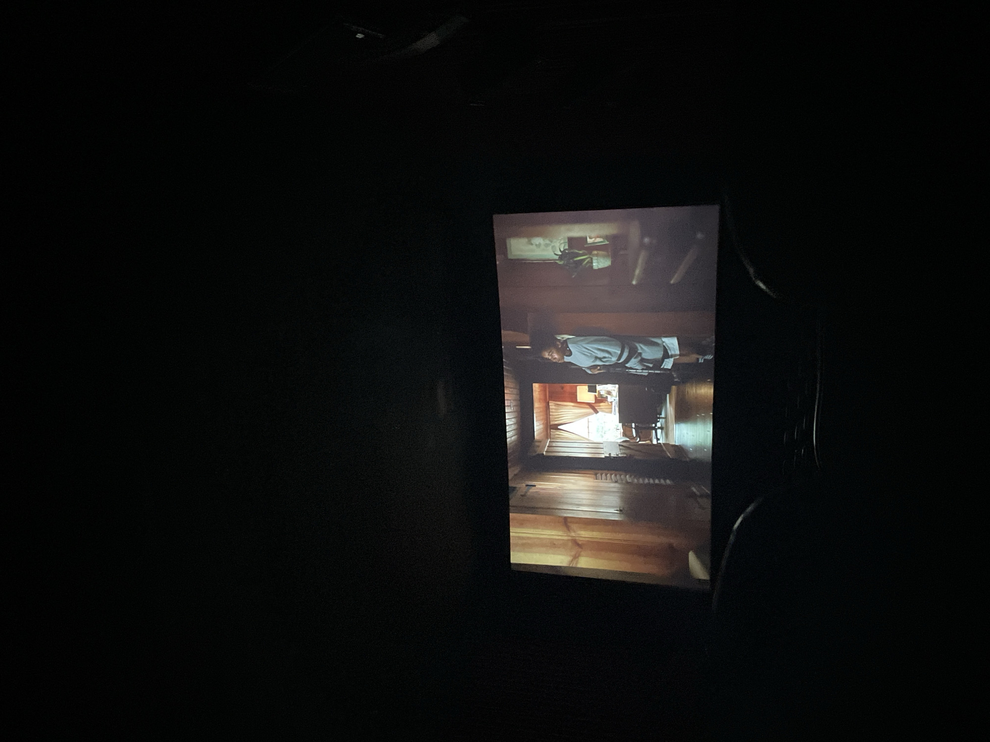 The image shows a darkened movie theater with a bright screen in the center. The screen depicts a scene inside a wooden building. In the scene, a person is lying on a cot. Buenos Aires, Argentina is not identifiable in the scene.