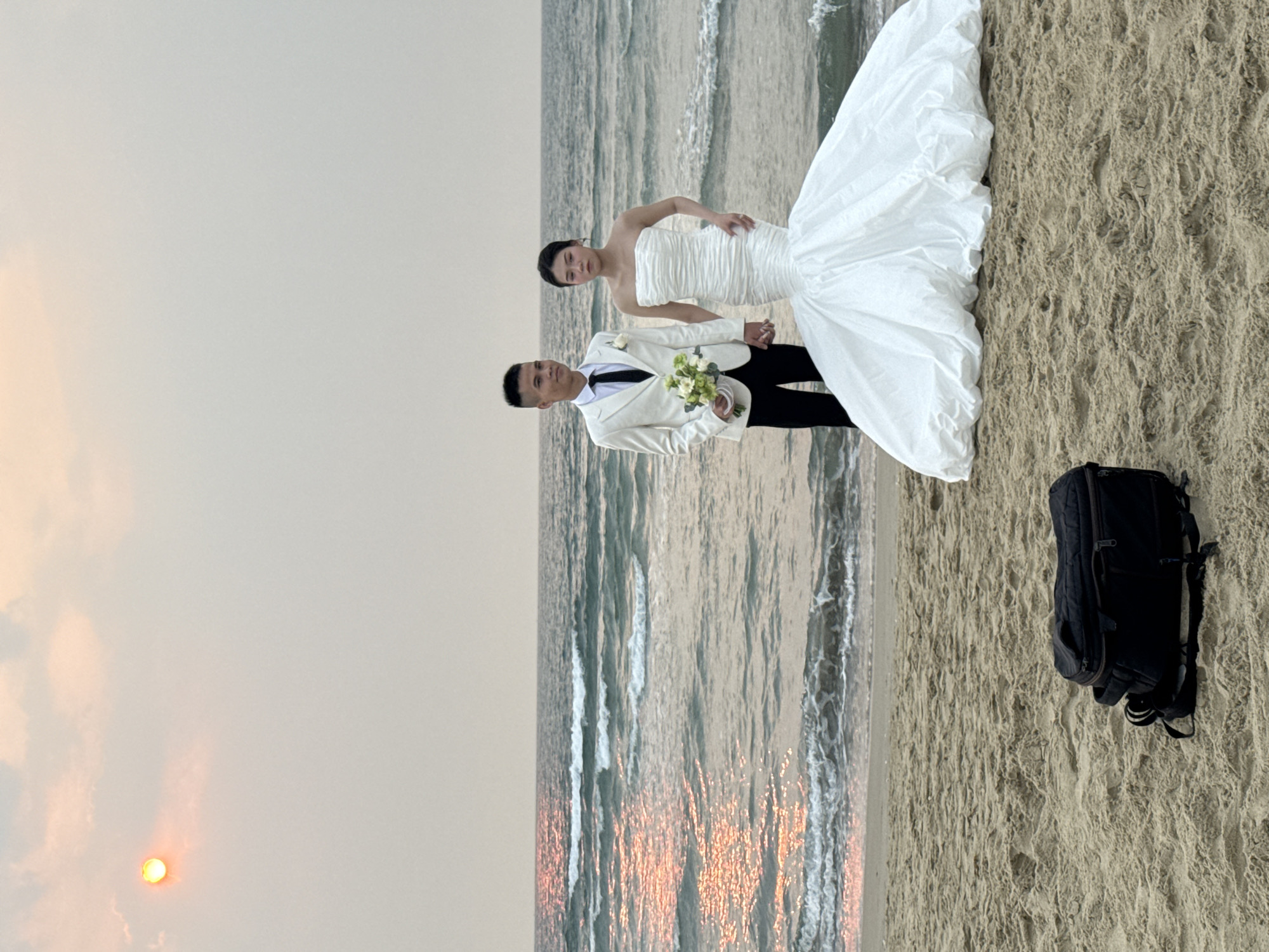A wedding scene unfolds on a beach in Đà Nẵng, Vietnam. A bride in a white, strapless wedding dress and a groom in a white suit and black pants stand hand-in-hand on the sand near the ocean. The bride has her left hand on her hip. The groom holds a bouquet of white and green flowers. The sun is setting, visible as a bright orange orb through the overcast sky. The ocean waves reflect the orange sunlight. A black backpack rests on the sand to the right of the couple.