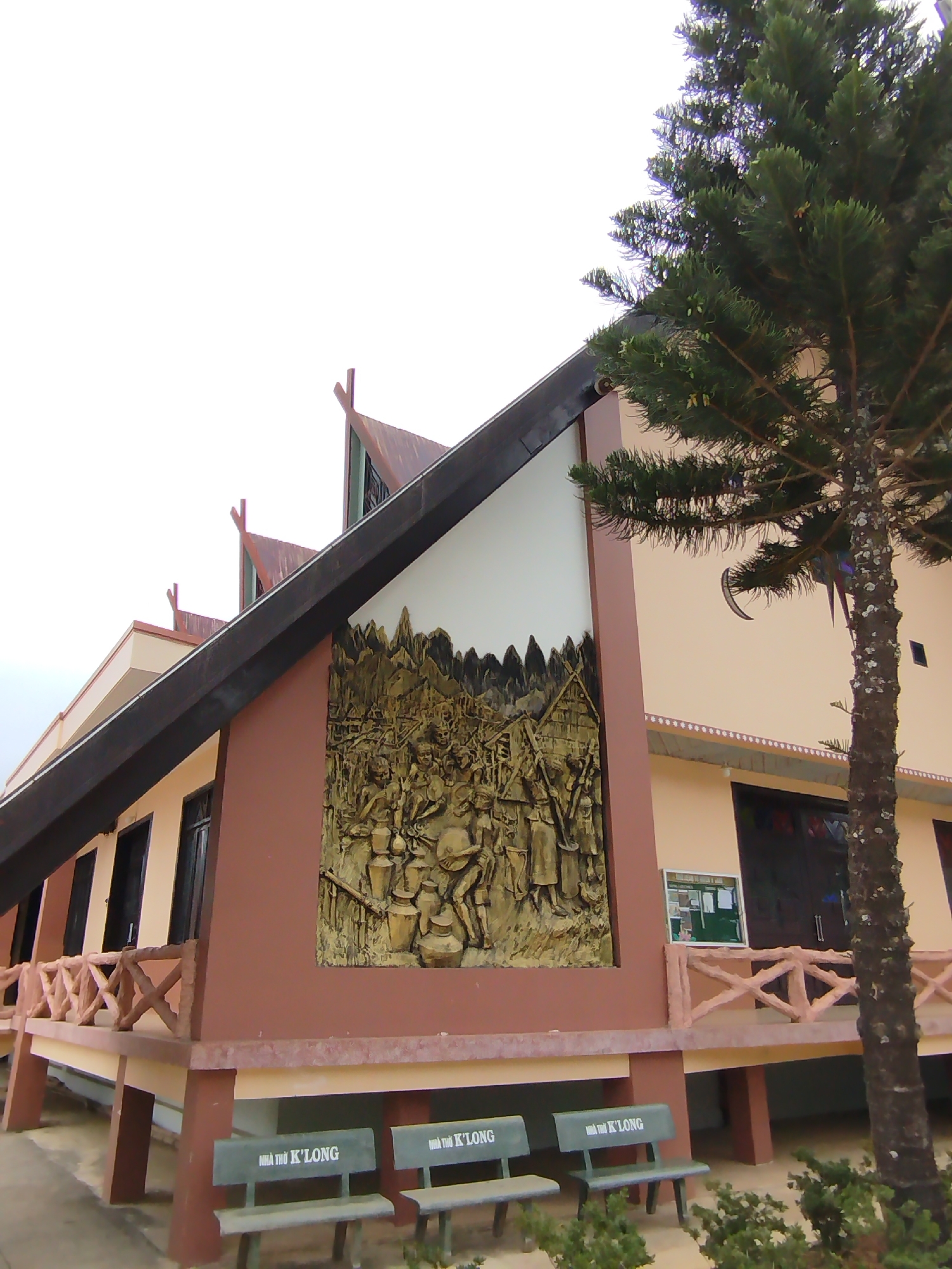 The image shows the facade of the Nhà Thờ K'Long church in K'Long, Vietnam. The building has a unique design with a triangular roof structure. A large, golden-colored bas-relief depicting a group of people in traditional attire is displayed on the building's exterior. Benches labeled "Nhà Thờ K'Long" are located in front of the building. A tree stands to the right of the building. A bulletin board is present. The sky is overcast.