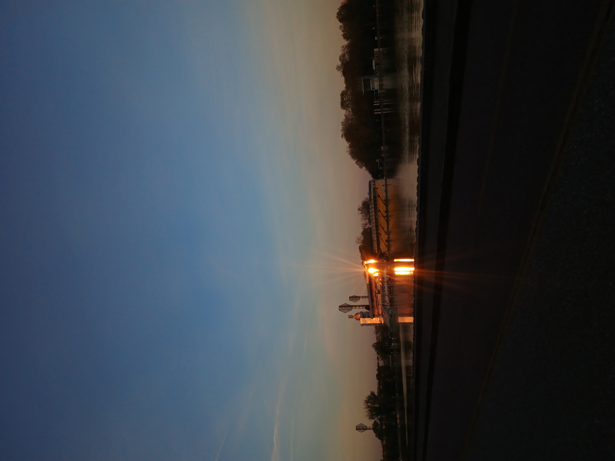 An outdoor scene captures a waterscape at twilight or night, with the image initially oriented sideways. When viewed correctly, a wide body of water spans the midground, reflecting the transitioning sky and artificial lights.
The sky, occupying the upper portion, displays a gradient from deep, clear blue at its zenith to lighter, warmer hues of orange and pink near the horizon, suggesting either a vivid sunset or early sunrise. No clouds are visible.
Across the water, on the distant bank, a line of trees forms a dark silhouette against the sky. Several prominent artificial light sources emit bright, star-like flares, casting intense golden reflections onto the calm water surface. These lights illuminate various structures on the bank, some appearing as decorative towers or potentially smaller Ferris wheel-like constructions. Buildings or other infrastructure are also faintly discernible behind the lights and trees.
In the immediate foreground, a dark surface, likely a road or paved pathway, features a faint yellow dividing line running horizontally. This foreground element is largely shadowed.
The scene conveys a peaceful and atmospheric ambiance, characteristic of a serene evening or early morning along a waterfront. No people are visible, and there is no discernible text. Given the context, this tranquil waterscape is likely located in Plovdiv, Bulgaria, possibly along the Maritsa River or a local park.