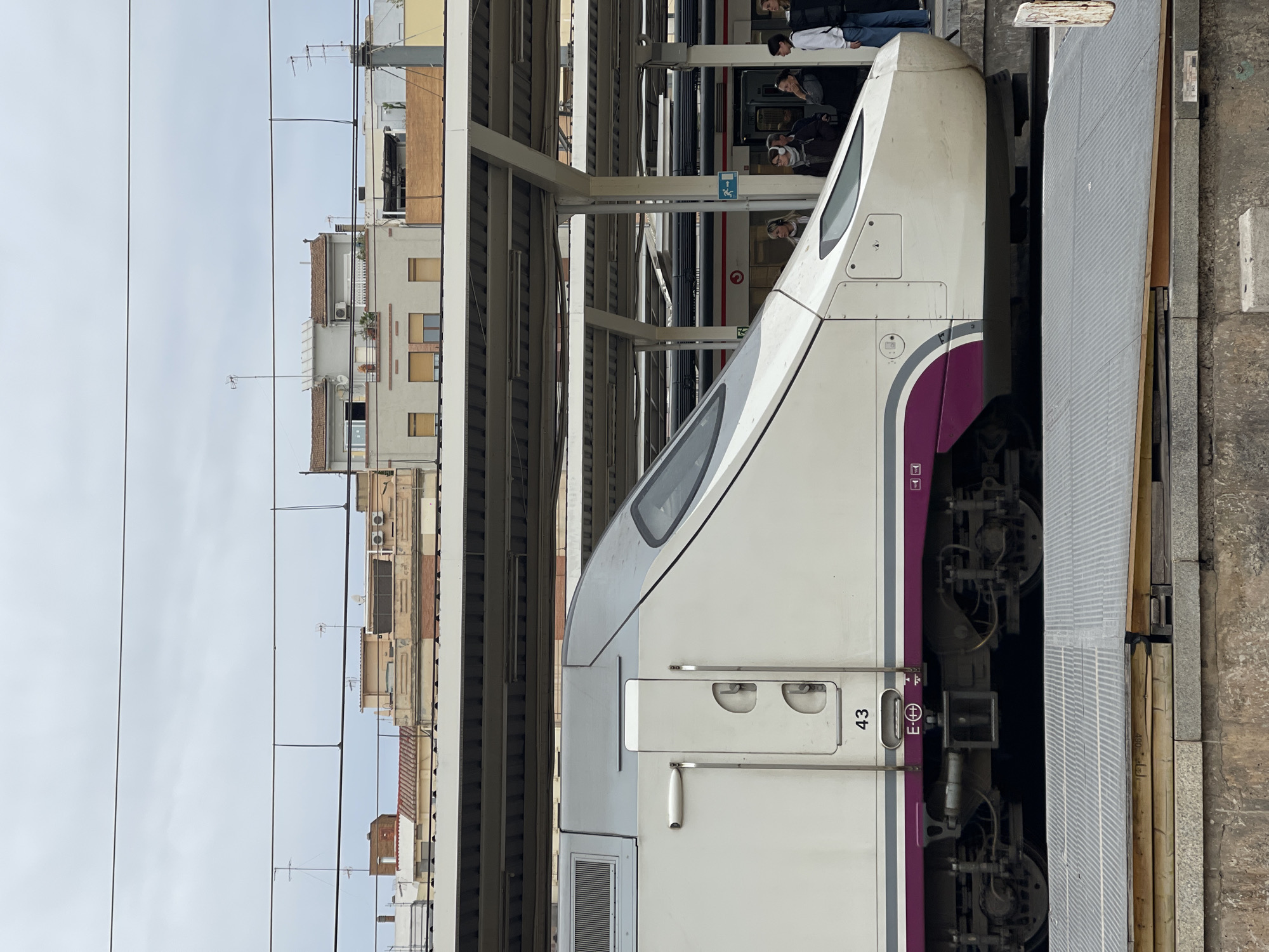 A close-up, angled shot depicts the front of a white and purple high-speed train at a station in València, Spain. Passengers are boarding or disembarking the train. "43" is visible on the side. Overhead structures cover the platform, while buildings are visible in the background. Overhead electrical lines are also present.