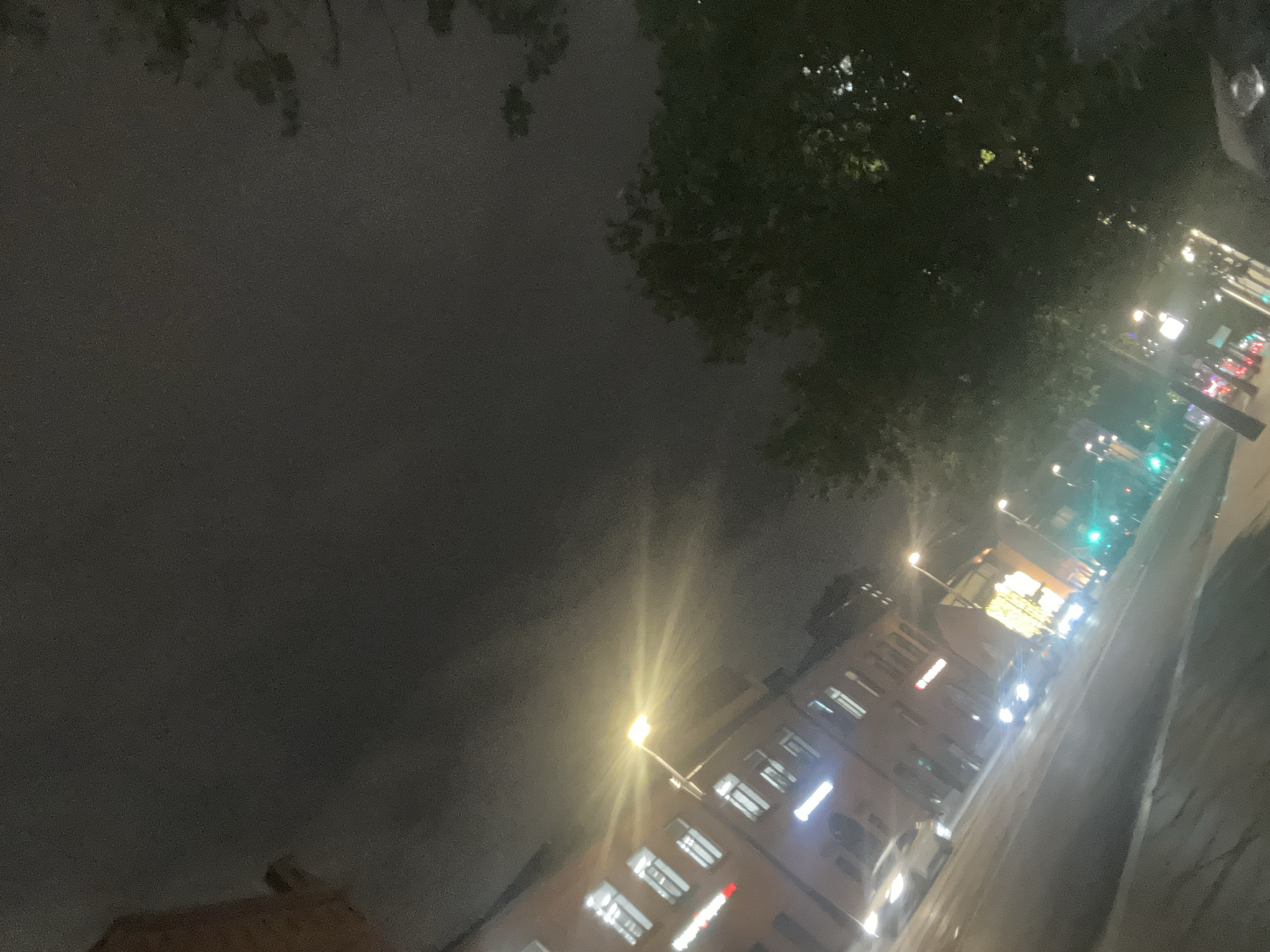 The image captures a nighttime urban scene, likely in Unknown, Unknown. A multi-story building with illuminated windows stands at an angle, with a streetlamp emitting a bright light next to it. Trees with dense foliage are seen above and to the side of the building. The sky is overcast and dark. A street with traffic lights, cars, and possibly pedestrians, runs along the right side of the image, obscured by the angle.