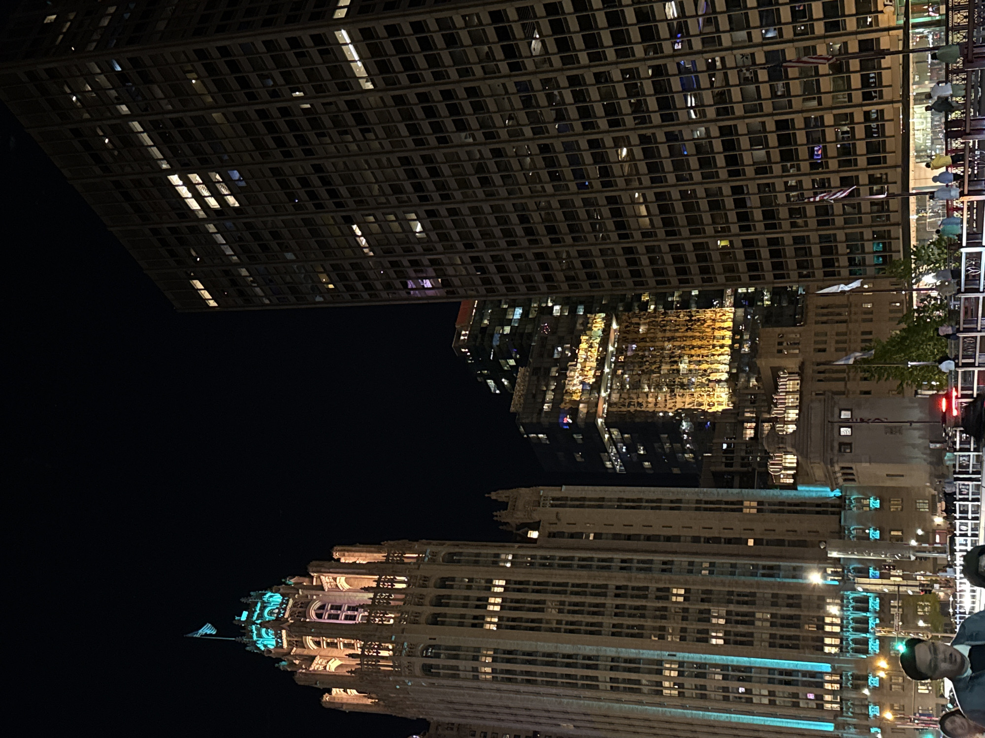 The image depicts a night scene in Chicago, United States. Prominent in the foreground is the Wrigley Building, recognizable by its illuminated, ornate neo-Gothic architecture. Rising behind it, the cityscape includes several other illuminated buildings, including a modern high-rise with a grid-like facade. Pedestrians and street furniture are visible on the right-hand side of the frame.