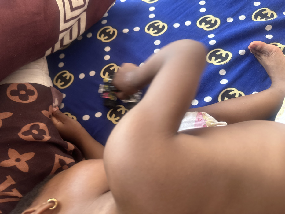 An overhead view depicts a young child, possibly a toddler, partially visible on a blue sheet with a golden interlocking "G" or "C" pattern and white polka dots, located in Iperu, Nigeria. The child's dark-skinned upper torso, right arm, and right leg are present in the frame. A single gold hoop earring is visible on the child's left ear. The child wears a white and pink patterned diaper or underwear. The right hand grasps a small, multi-colored toy, possibly a vehicle, with black and yellow details. The child's head and left side are partially obscured by a brown pillow featuring a repeated stylized quatrefoil and star pattern. Another brown pillow with a white and brown geometric pattern borders the scene on the right. The setting appears to be an indoor space, likely a bed.