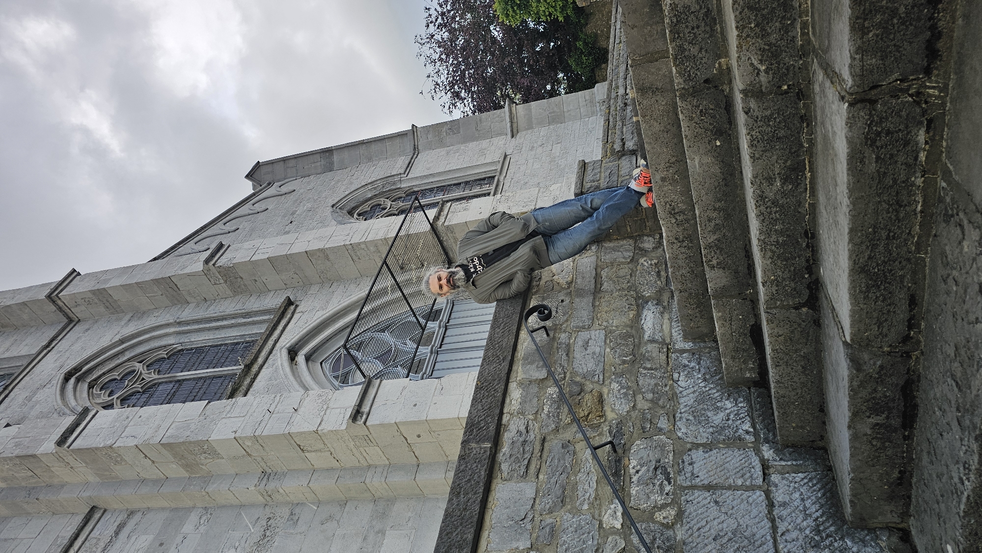 An outdoor portrait photograph shows a man standing on a stone-paved ground next to a large stone building and a set of stone steps. The man, who has grey hair and a beard, wears a grey jacket, blue jeans, and red and white sneakers. He is facing the camera with a slight smile.
The building, to the man's left, is constructed from light grey stone and features Gothic-style architecture. It has several ornate windows, including a large arched window with intricate tracery. The building's roofline shows decorative elements against an overcast sky. Patches of green and reddish-brown foliage are visible above the building.
To the man's right, dark grey stone steps ascend upwards, flanked by a stone wall with a black metal handrail. The ground he stands on is paved with irregular stone blocks. Text partially visible on his jacket appears to resemble "adidas". The scene takes place during the day under cloudy weather, characteristic of a historic European location such as Boussu-lez-Walcourt, Belgium.