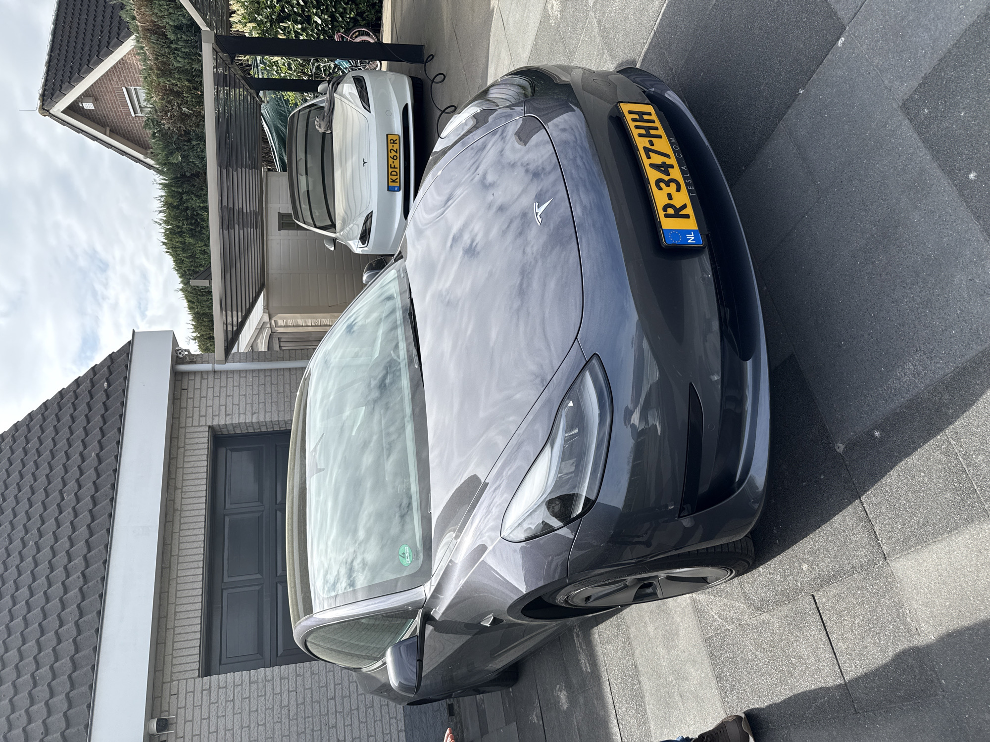 Here is a description of the image:
The image shows two Tesla cars parked on a paved driveway in front of a building. The car in the foreground is dark gray, while the one in the background is white. Both cars have Dutch license plates, with the gray car's plate reading "R-347-HH" and the white car's plate reading "KDF-62-R". The cars are parked in what appears to be a residential area, with a house and garden visible in the background. The focus of the image is on the front of the gray Tesla and its license plate.