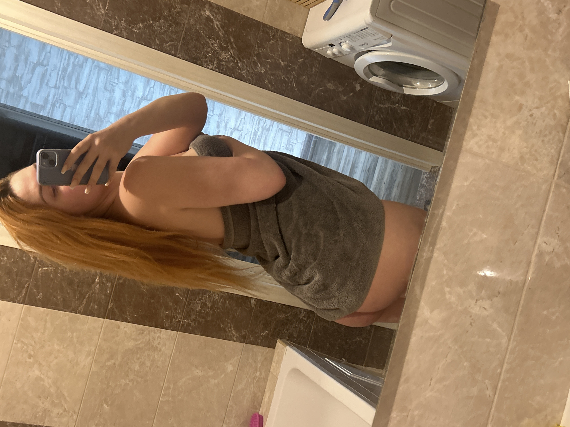 In a bathroom with tiled walls and a marble countertop, a woman with long, red hair is seen from the side, taking a selfie in a mirror. She is wrapped in a gray towel, covering her body from her chest down. Her right hand holds a smartphone, which is reflecting the image. A white washing machine is visible in the background to the right of the woman. A white bathtub is partially visible on the left side of the frame.