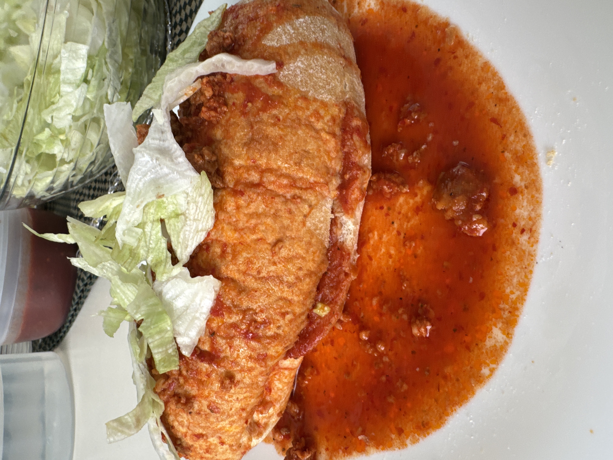 A close-up shot shows a pambazo, a Mexican sandwich, on a white plate. The pambazo is filled with chorizo and potatoes, topped with shredded lettuce, and partly submerged in a pool of red salsa. A container of shredded lettuce sits adjacent to the sandwich, partially visible in the upper left corner. Another small container, seemingly containing salsa, is located next to the lettuce container. The background includes a glimpse of a black and white patterned surface. There are no people visible in the image. The location is identified as San Bernardino Tlaxcalancingo, Mexico.