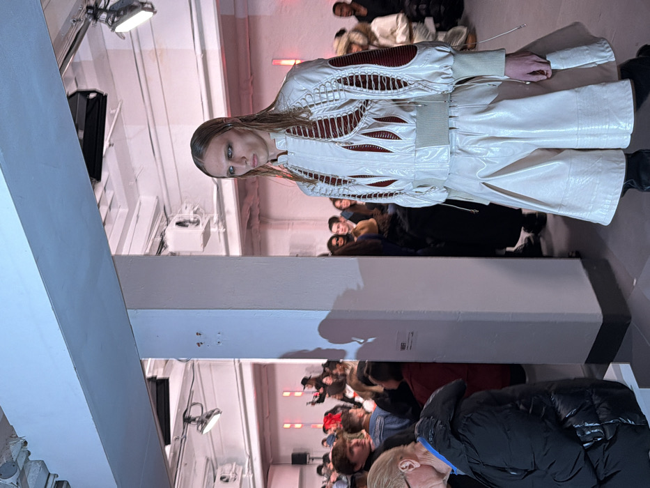 A fashion model showcases a white, futuristic-looking outfit with cut-out detailing on a runway. The model stands in front of an audience seated along the sides of the runway. Industrial elements, such as lighting fixtures, ceiling structures, and a large white pillar, are visible in the background. The setting suggests a fashion show in an urban or warehouse-style location.