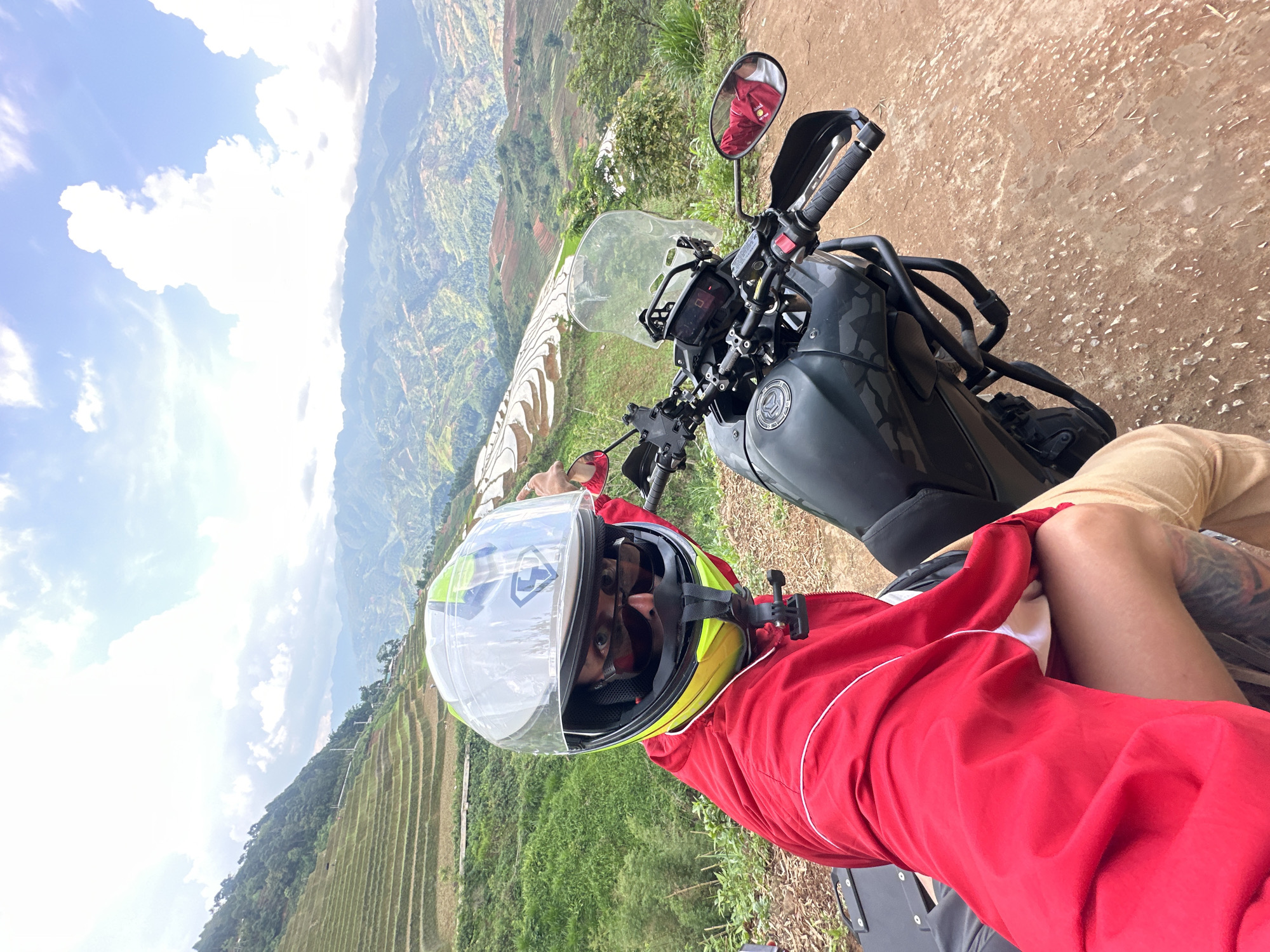 A first-person perspective image captures a person in a red jacket and a yellow and green helmet, seated on a black adventure motorcycle. The motorcycle is parked on a reddish-brown dirt path, with its handlebars, windshield, and a camouflaged fuel tank visible. An action camera is mounted on the helmet. The person's eyes are visible through the helmet's clear visor, looking directly at the camera. A tattoo is visible on their right arm or leg.
The scene unfolds in a mountainous region, characteristic of Lao Chai, Vietnam. In the midground, vast and intricate rice terraces, some appearing freshly cultivated and others lush green, carve their way down steep hillsides. Beyond these agricultural steps, distant mountains stretch into a hazy horizon, crowned by a bright blue sky dotted with fluffy white clouds. The weather is clear and sunny, suggesting it is daytime. The image portrays a moment of rest during a motorcycle journey, pausing at a scenic overlook to appreciate the breathtaking landscape. No discernible text is clearly visible on the helmet or motorcycle.