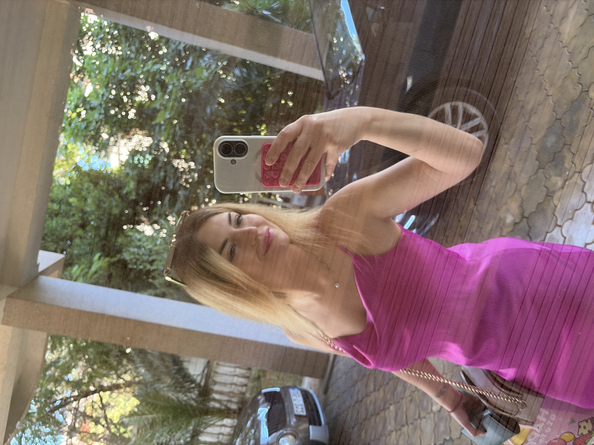 A woman takes a selfie in a reflective surface, likely a window or a glass door, of a building in Budva, Montenegro. She is wearing a bright pink, sleeveless, cowl-neck dress with a ribbed texture and delicate gold chain straps. She holds a white smartphone with a silver case and a pink grip attachment in her right hand, angled to capture her reflection. A delicate silver necklace with a small pendant is visible on her neck. Her blonde hair is parted to the side and falls loosely around her shoulders. She wears sunglasses pushed up on top of her head. In the background, seen through the reflective surface, are lush green trees with sunlight filtering through, suggesting an outdoor setting. To the left, the front of a dark-colored car is partially visible, parked on a paved driveway. The ground appears to be paved with interlocking stones. The overall impression is a casual, outdoor moment captured by the woman.