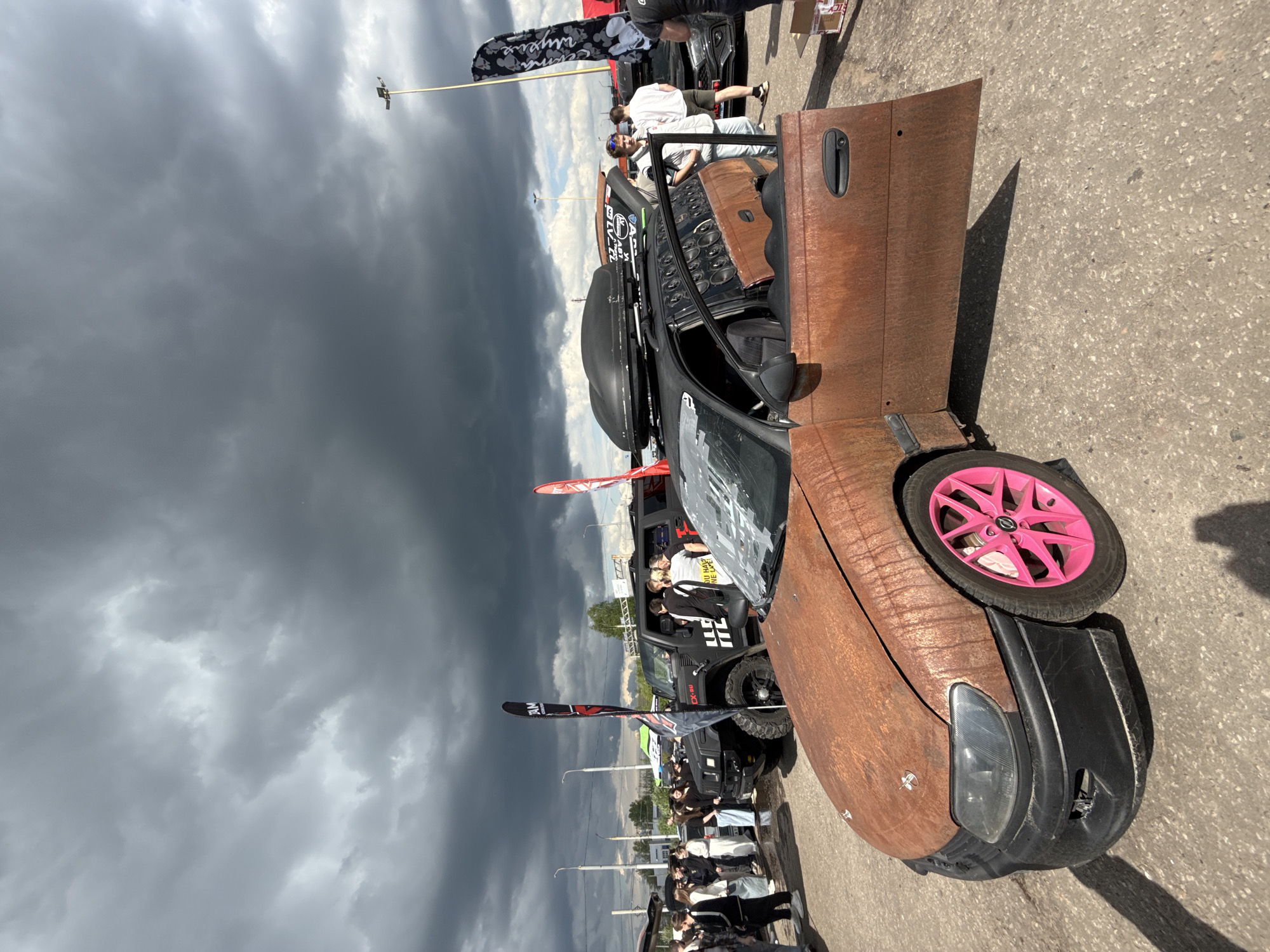 The image shows a rusted car with bright pink wheels and an open door at an event with a large crowd under a stormy sky. There are other cars and advertising banners visible in the background. The location is Unknown, Unknown.
